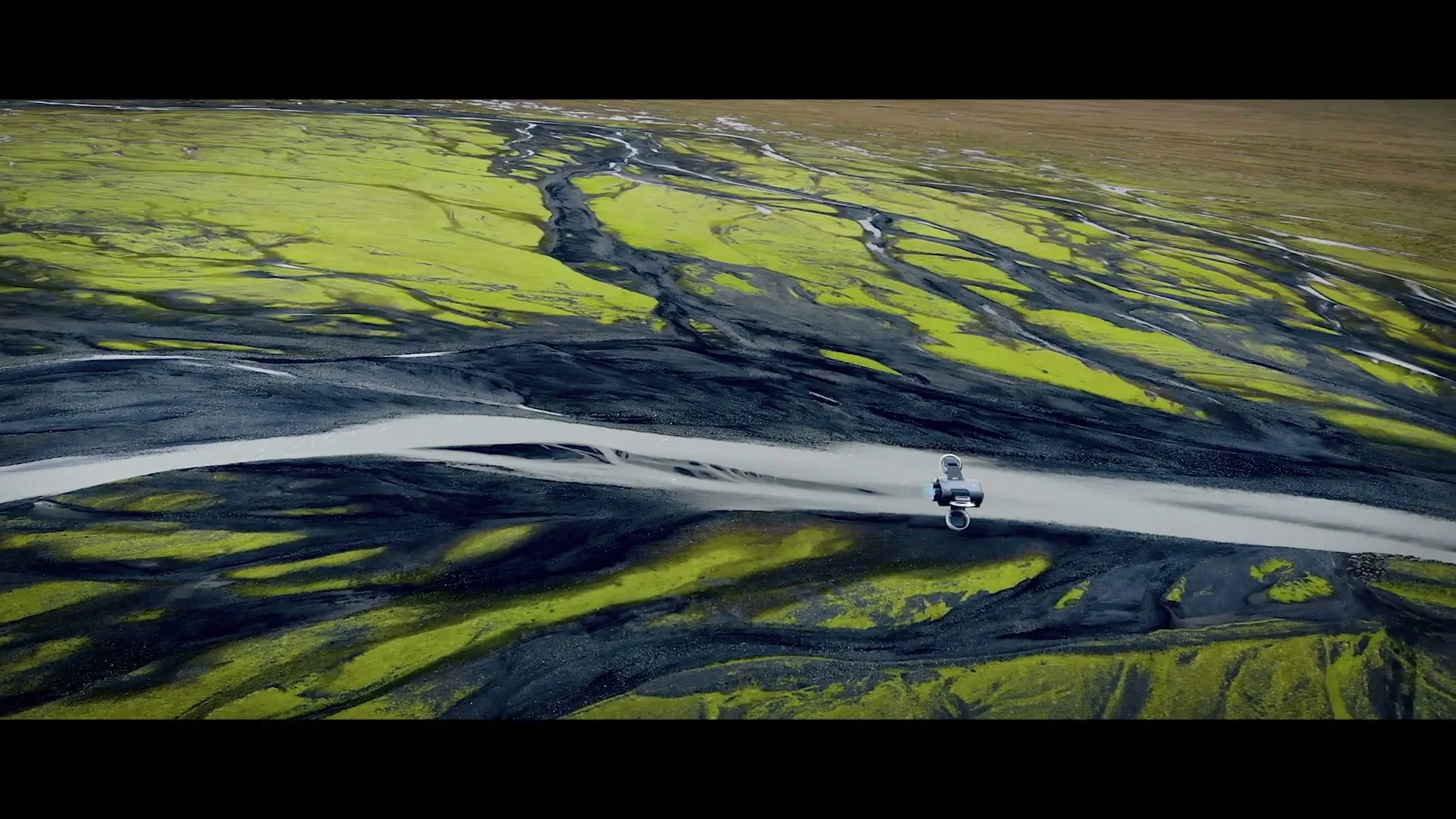 An astronaut in a white spacesuit stands on a narrow strip of land between two rivers, surrounded by vibrant green moss and dark volcanic rock.