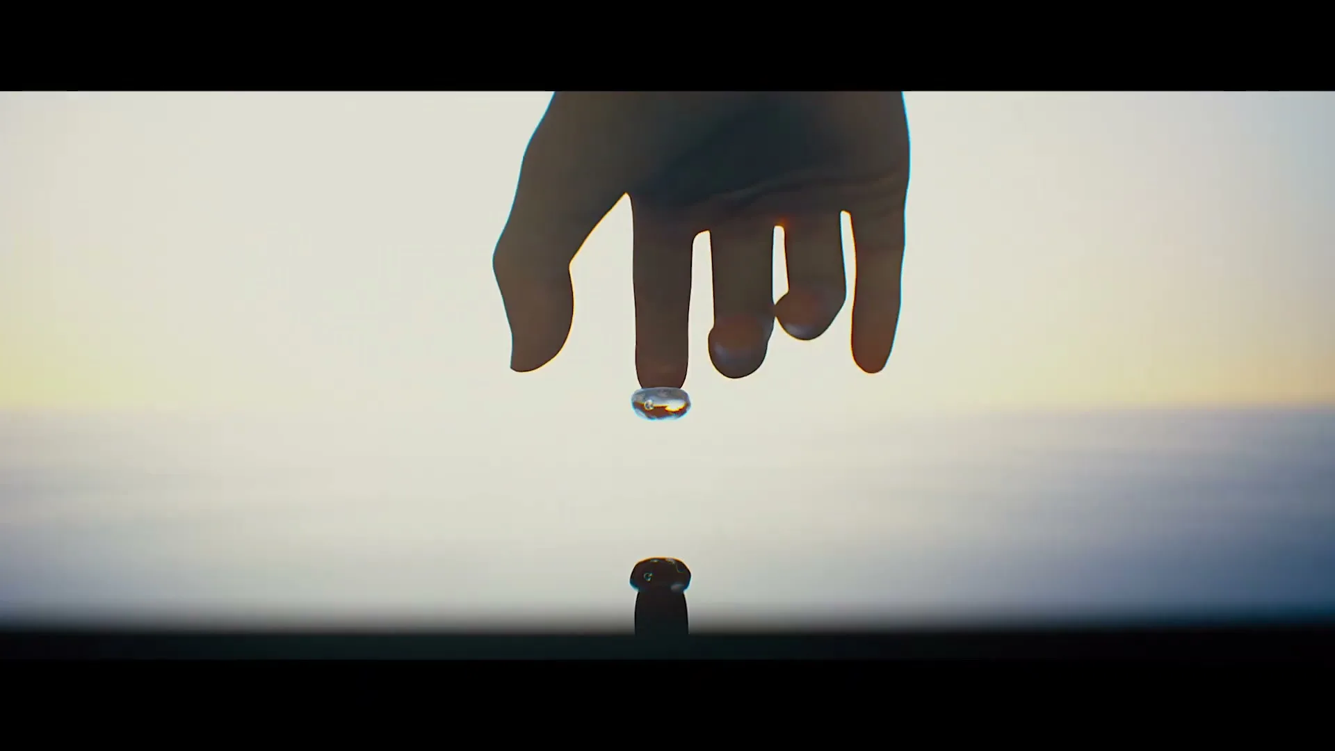A hand with a water droplet on its finger, hovering over another droplet on a surface, against a gradient sky.