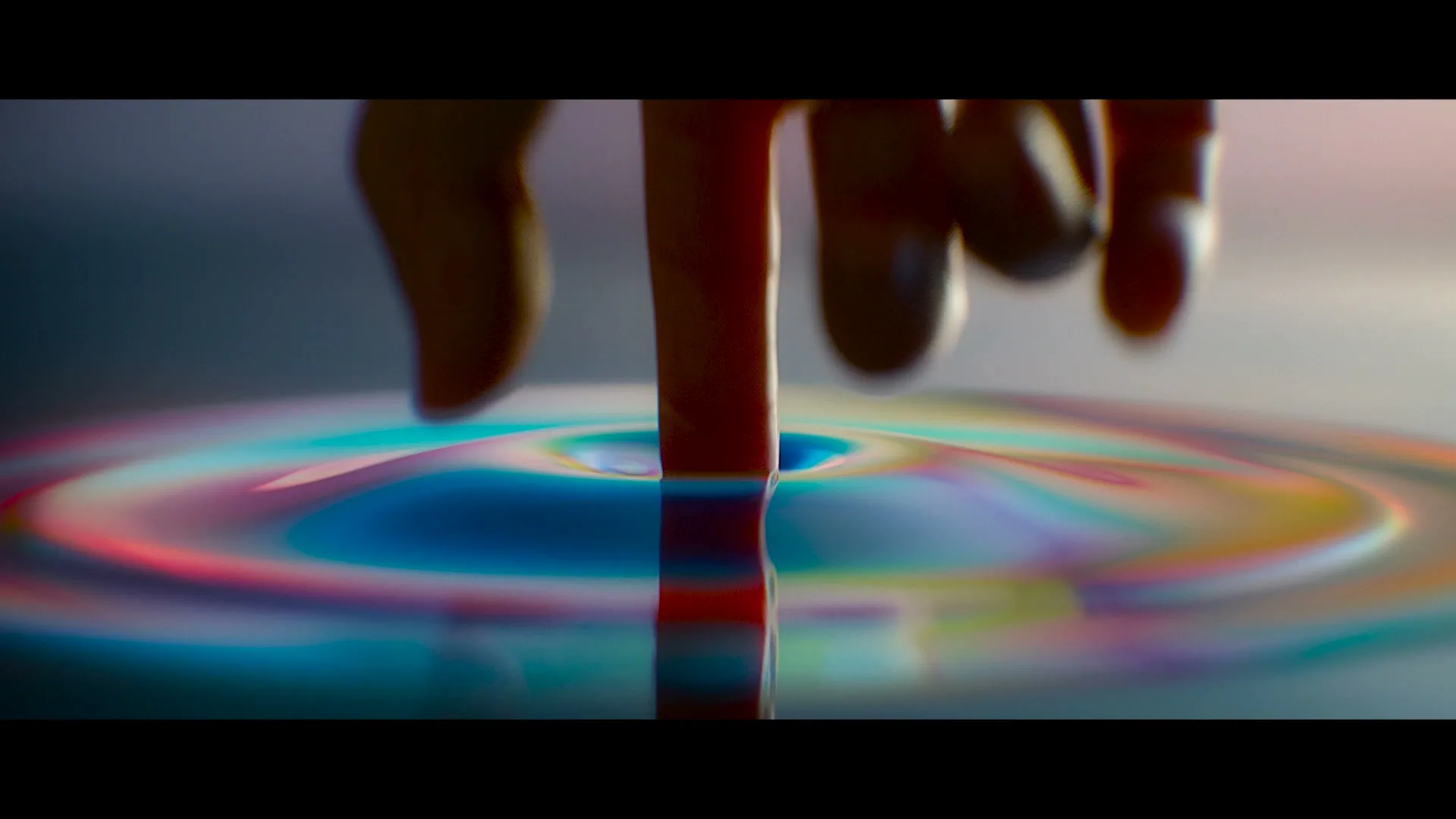 A human finger touches a liquid surface, creating colorful ripples in a studio setting.