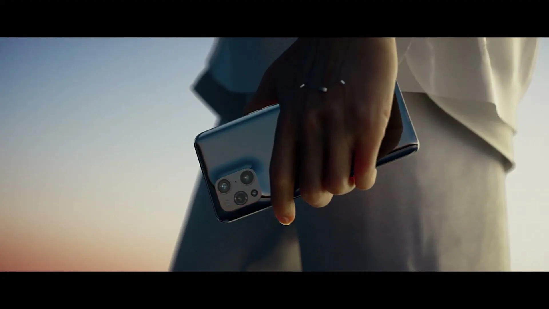 A person in white clothing holds a black smartphone with a unique camera bump, walking against a soft, colorful sky at sunset.
