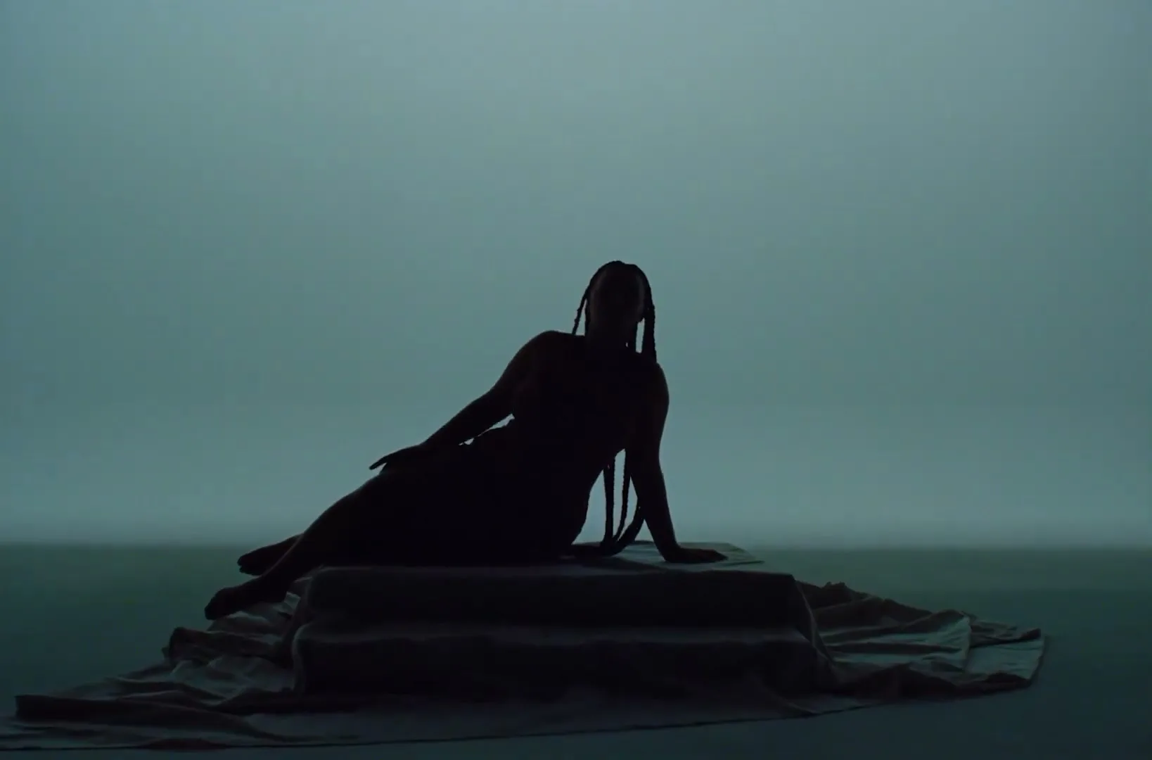 A woman with long braids in a red and blue dress reclines on a draped platform in a studio, transitioning from silhouette to full visibility.