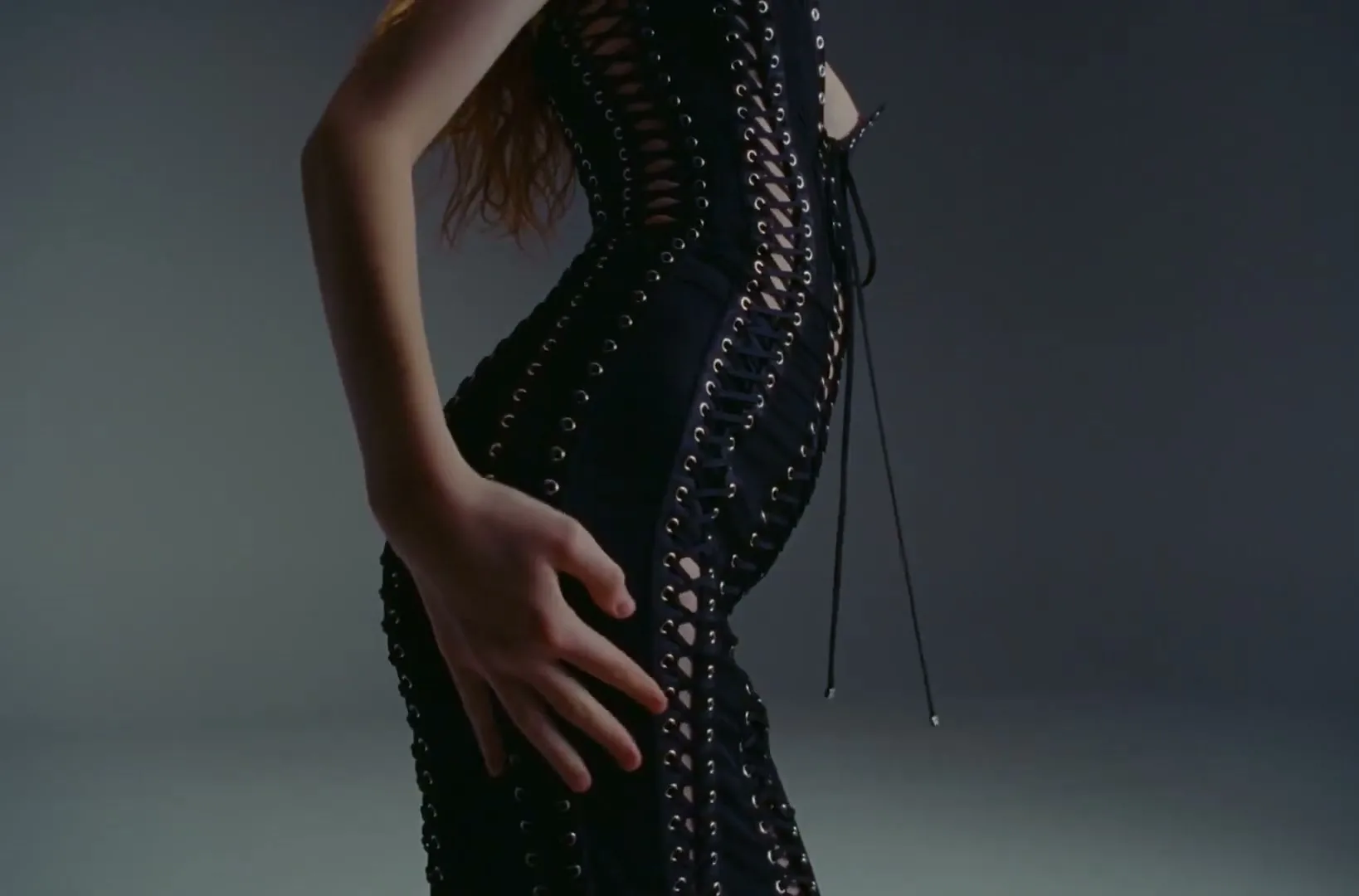 A woman with long red hair, wearing a black lace-up dress, poses in a studio, touching her hip.