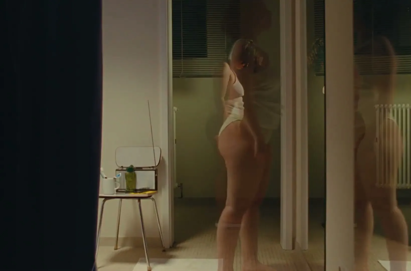 A woman with short curly hair, wearing a white tank top and light-colored underwear, stands on a scale in a bathroom, looking down at her feet.