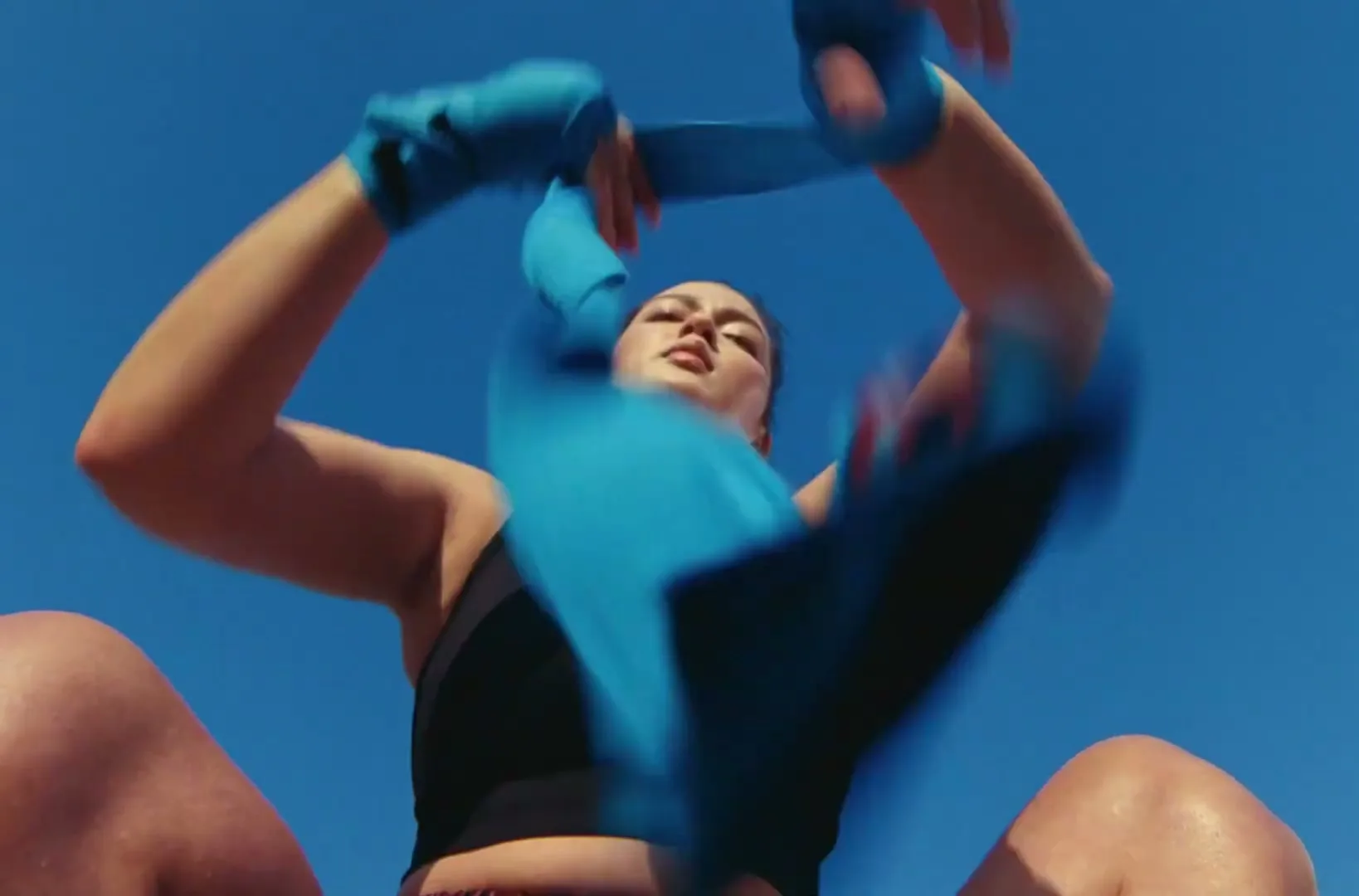 A woman with dark hair and a black sports bra wraps blue boxing tape around her hands, looking focused against a clear blue sky.
