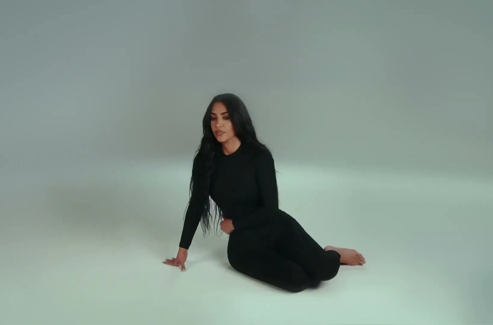 Kim Kardashian, a woman with long dark hair, wearing a black long-sleeved top and pants, sits on the floor in a studio, looking to the side.