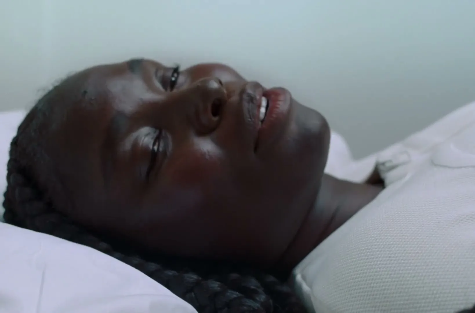 A young Black woman with braided hair lies on a white pillow, smiling and talking, in a bright, minimalist room.