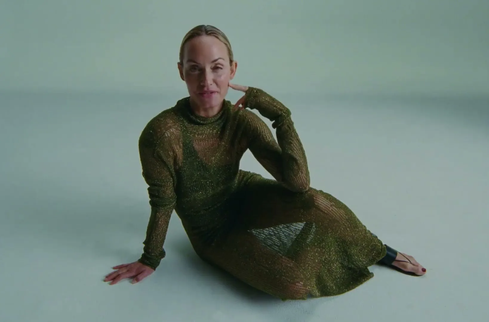 A woman in her 40s, wearing a sheer green dress, sits on the floor in a studio, looking at the camera and talking.