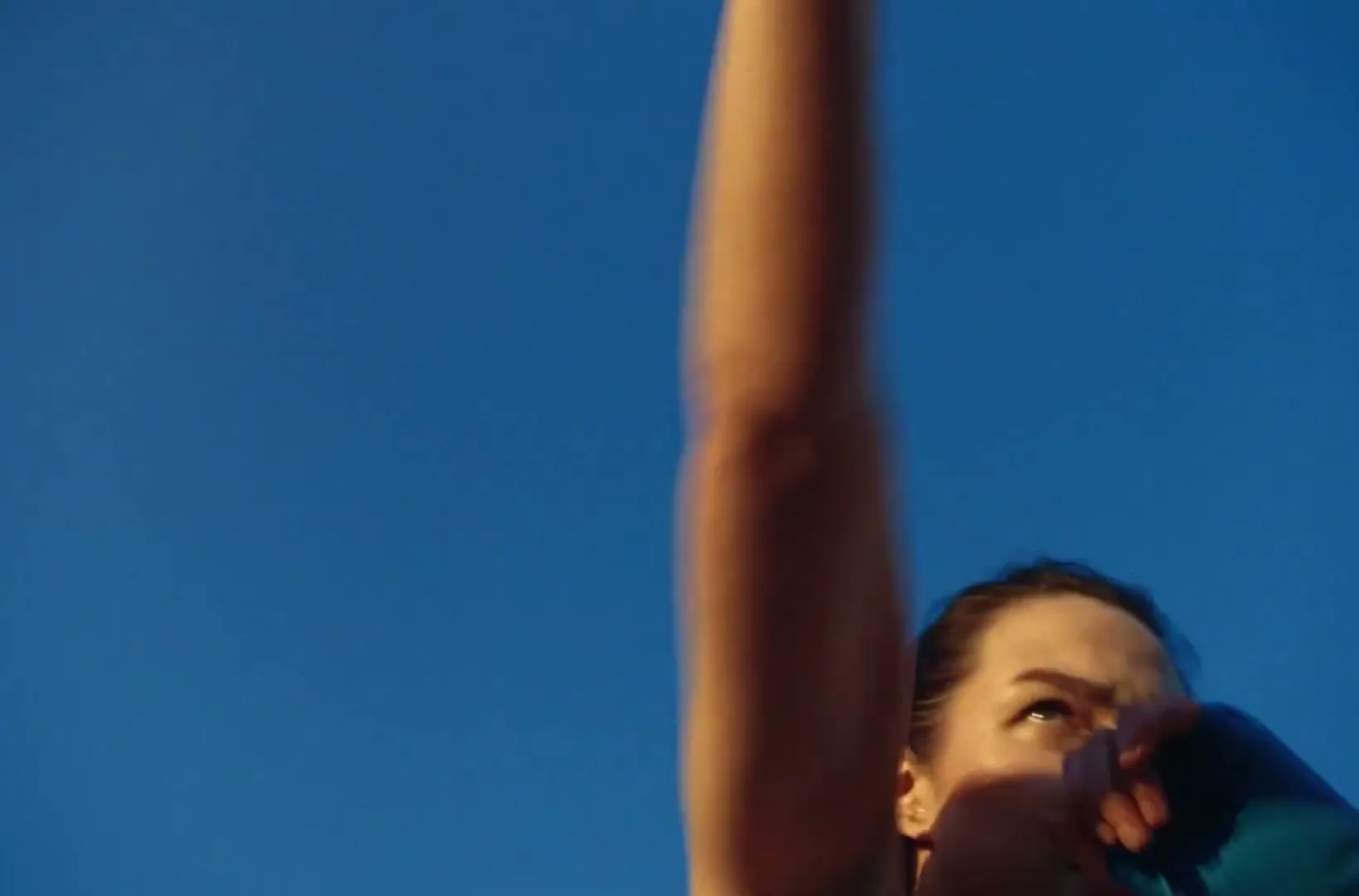 A woman in her 20s, wearing a teal top, is seen from a low angle against a clear blue sky, looking up with a determined expression.