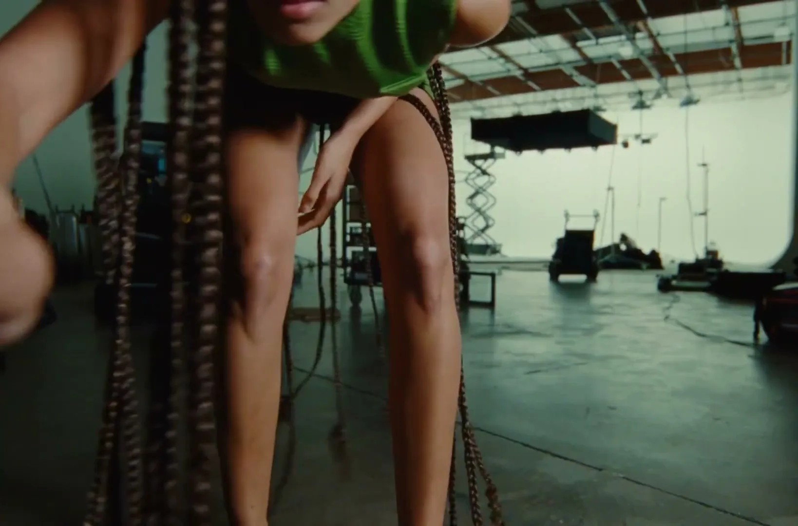 A young woman with long braids, wearing a green top and shorts, bends down in a large, industrial studio space.