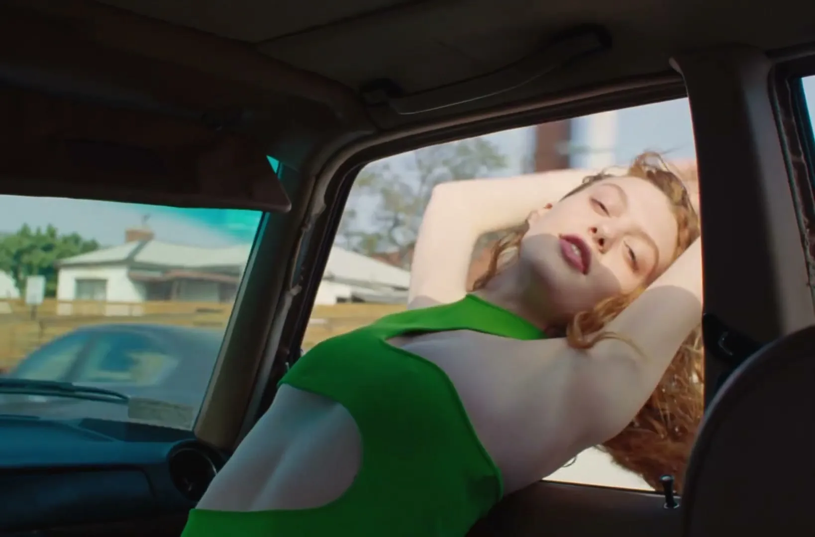 A young woman with red hair, wearing a green cutout top, leans out of a car window, stretching her arms above her head, on a sunny day.