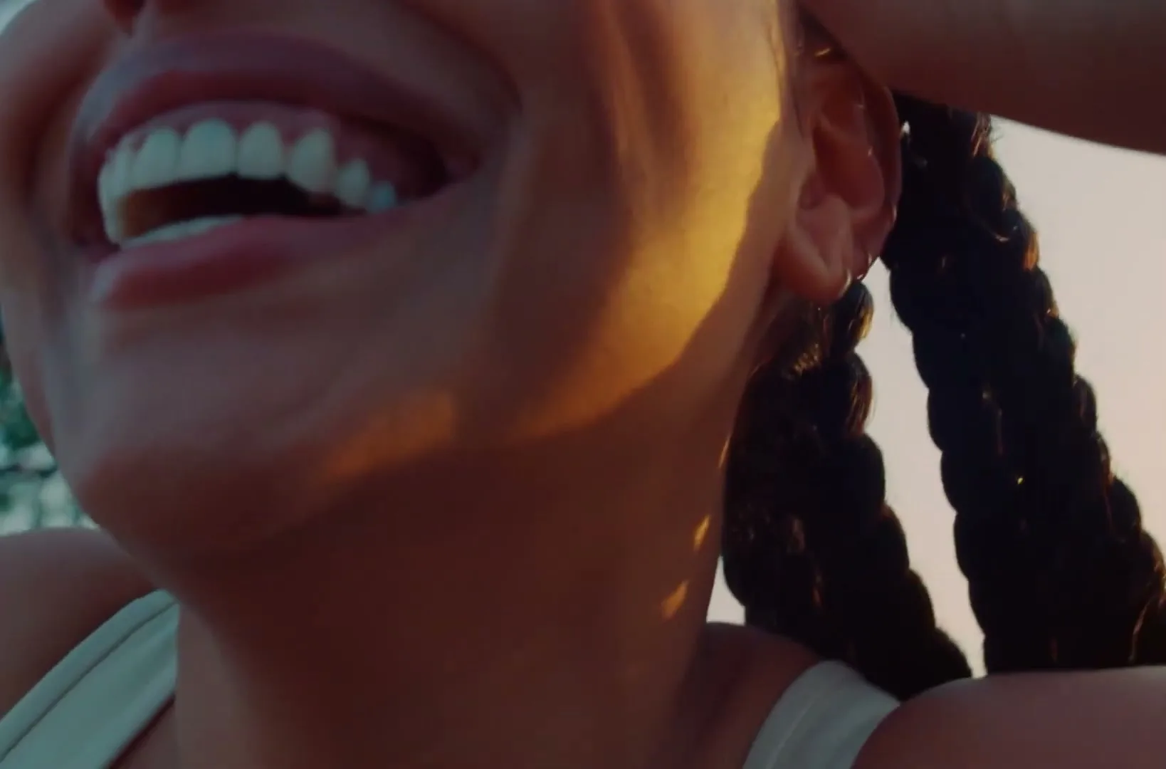 A woman with braided hair laughs joyfully, her mouth open, with golden hour light on her face in an outdoor setting.