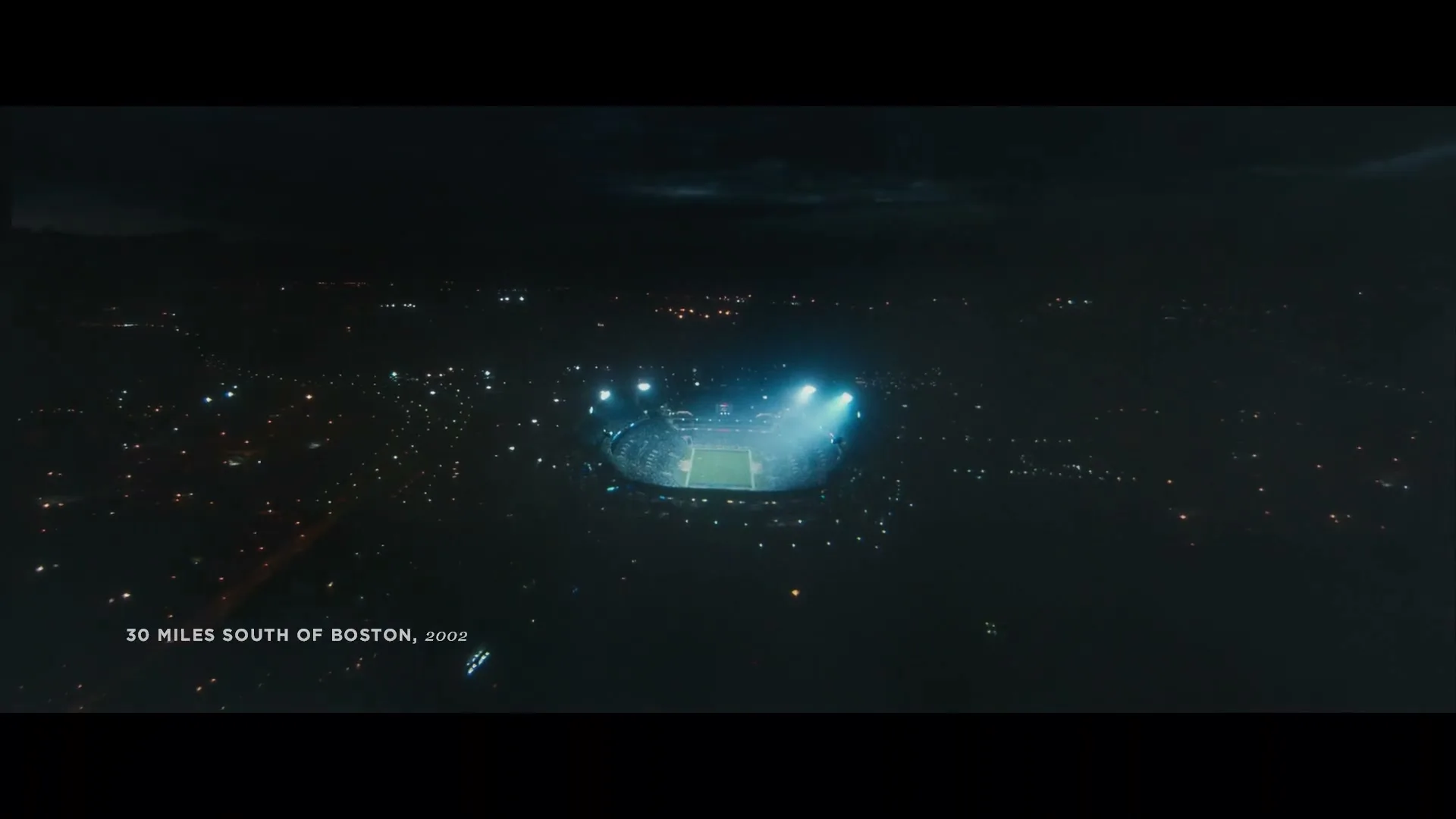An aerial view of a brightly lit stadium at night, surrounded by the dark expanse of a city with scattered lights, with text on screen.