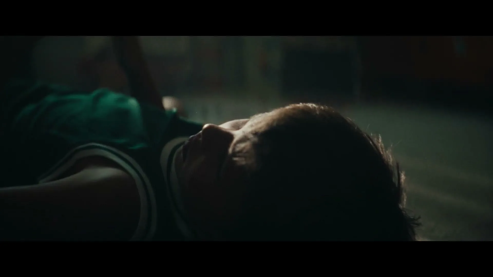 A young male basketball player lies on the court, looking up, with a basketball resting on his chest in a dimly lit gym.