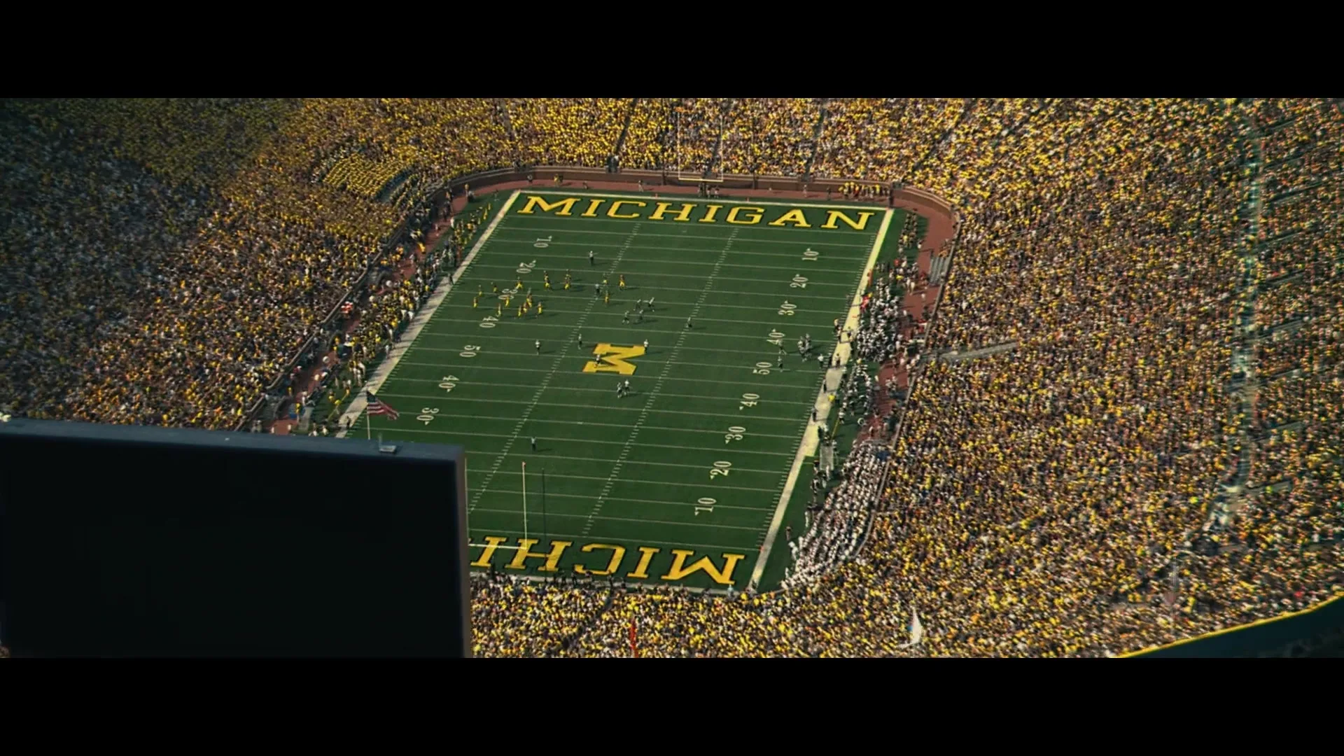 An aerial view of a packed football stadium, with a game in progress on the green field, surrounded by thousands of cheering fans.
