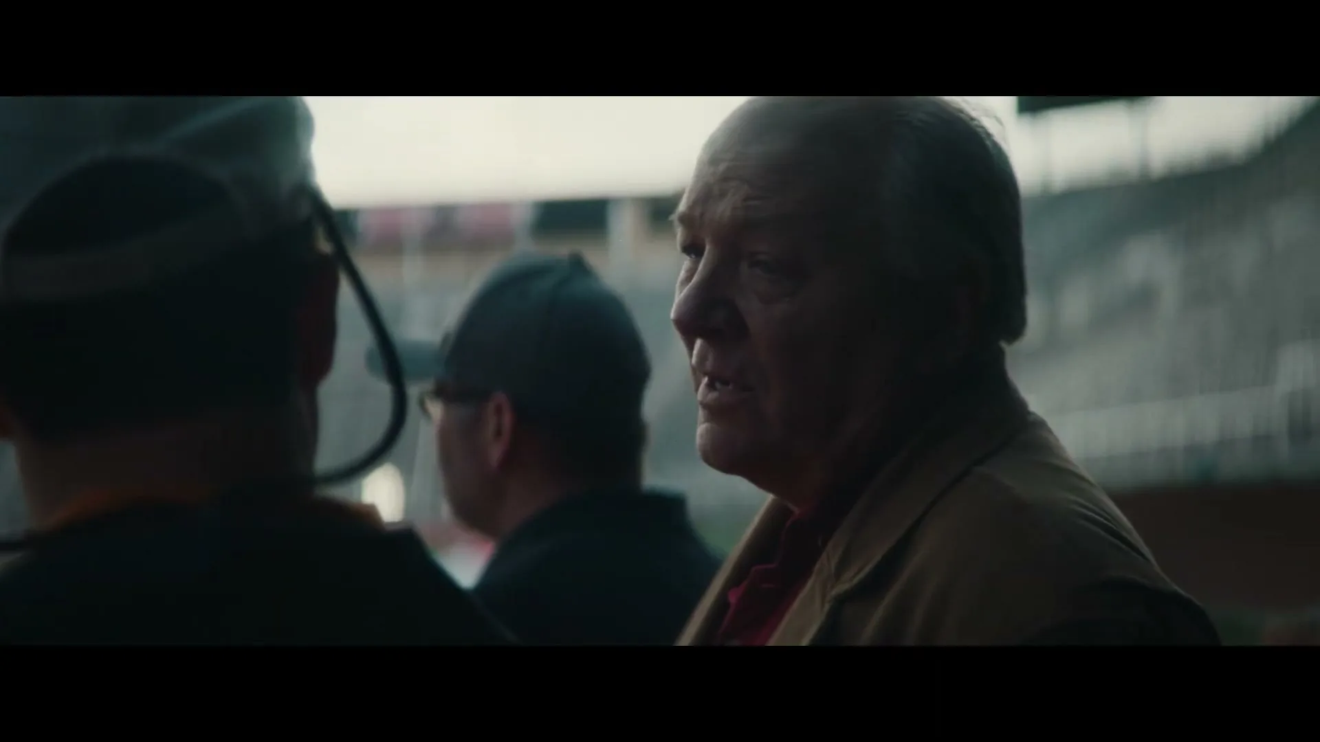 An older man in a brown jacket and red shirt looks to the right, talking to two other men in a stadium.