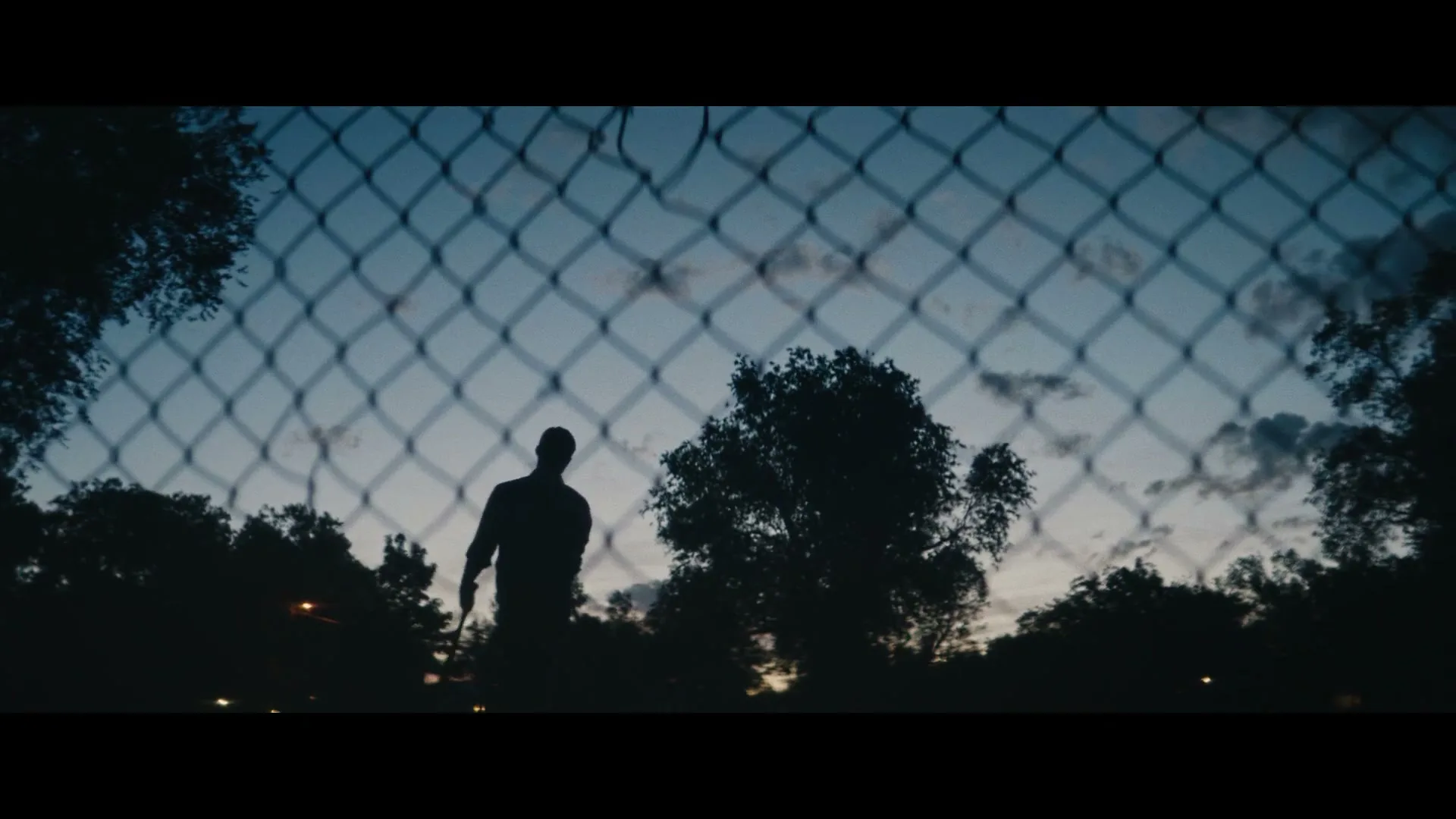 A silhouetted male baseball player swings a bat, seen through a chain-link fence at dusk in a park.