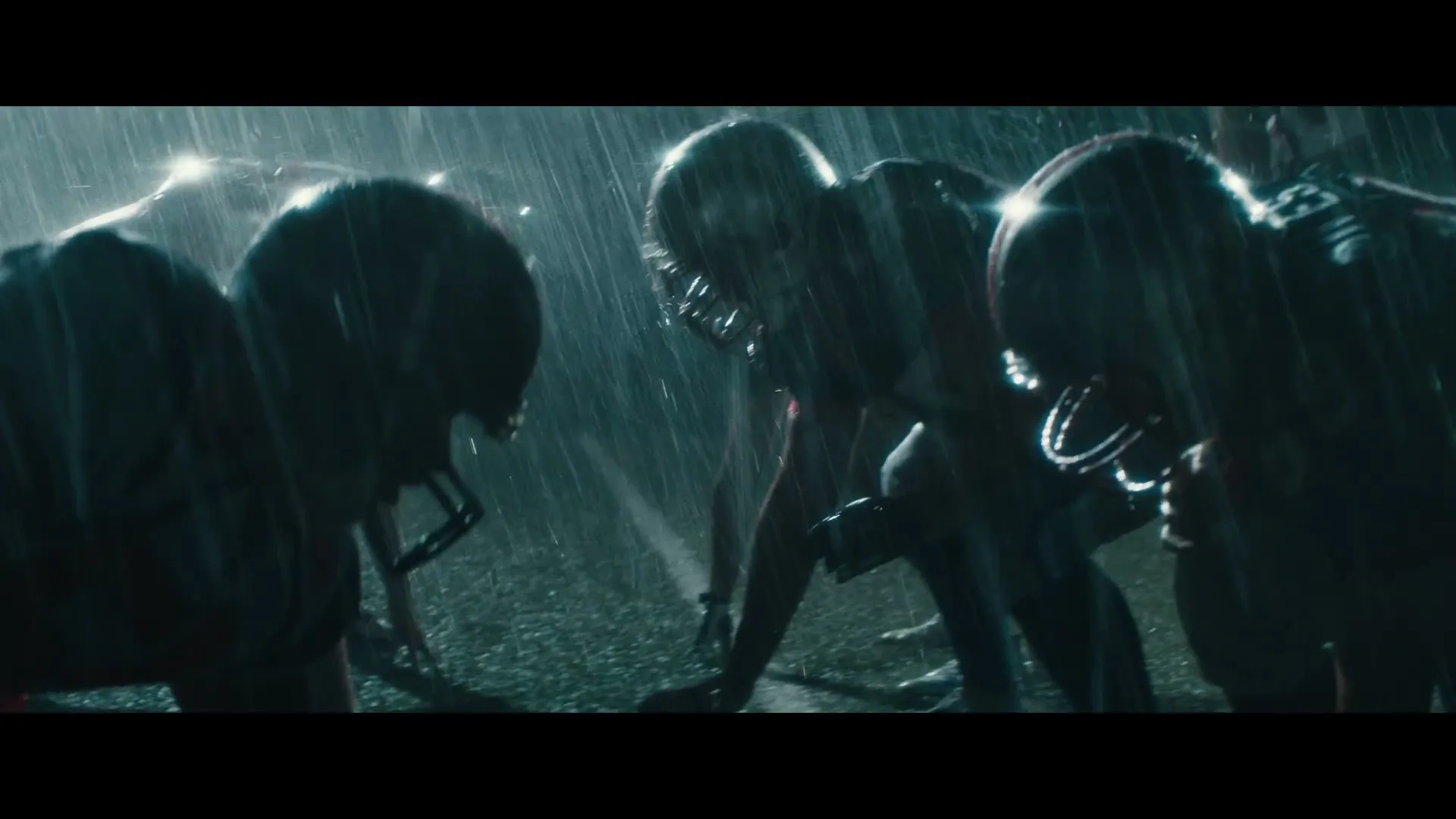 American football players in helmets and pads crouch on a wet field, ready to play in the rain at night.