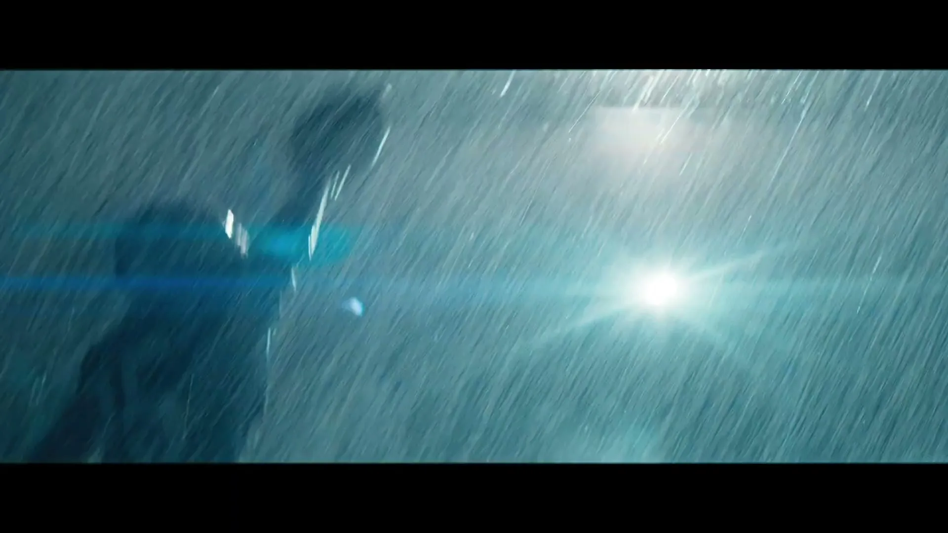 A man in a dark suit stands in heavy rain, illuminated by a bright light source, creating a dramatic and intense atmosphere.