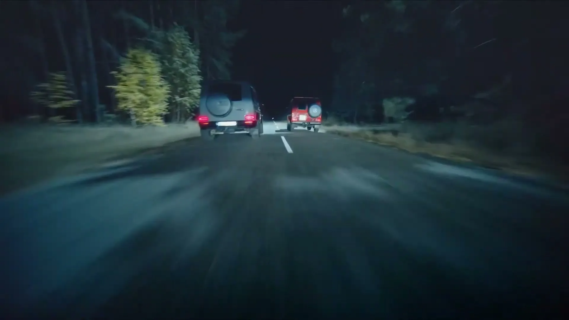 Two Mercedes-Benz G-Class SUVs, one grey and one red, drive down a dark, tree-lined road at night, with the camera following closely behind the grey vehicle.