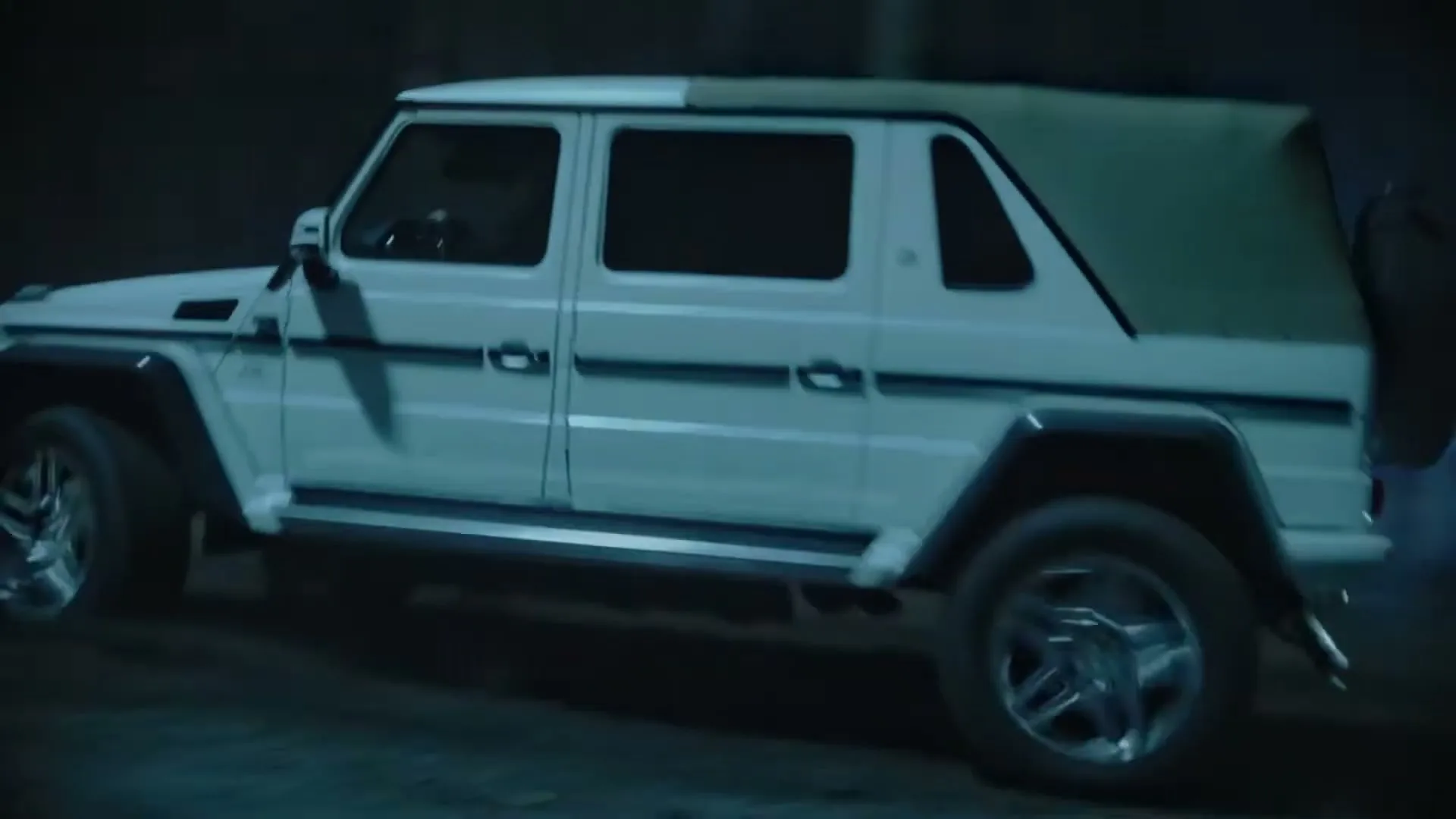 A white Mercedes-Benz G-Class SUV drives at night on a dirt road, with its headlights on.