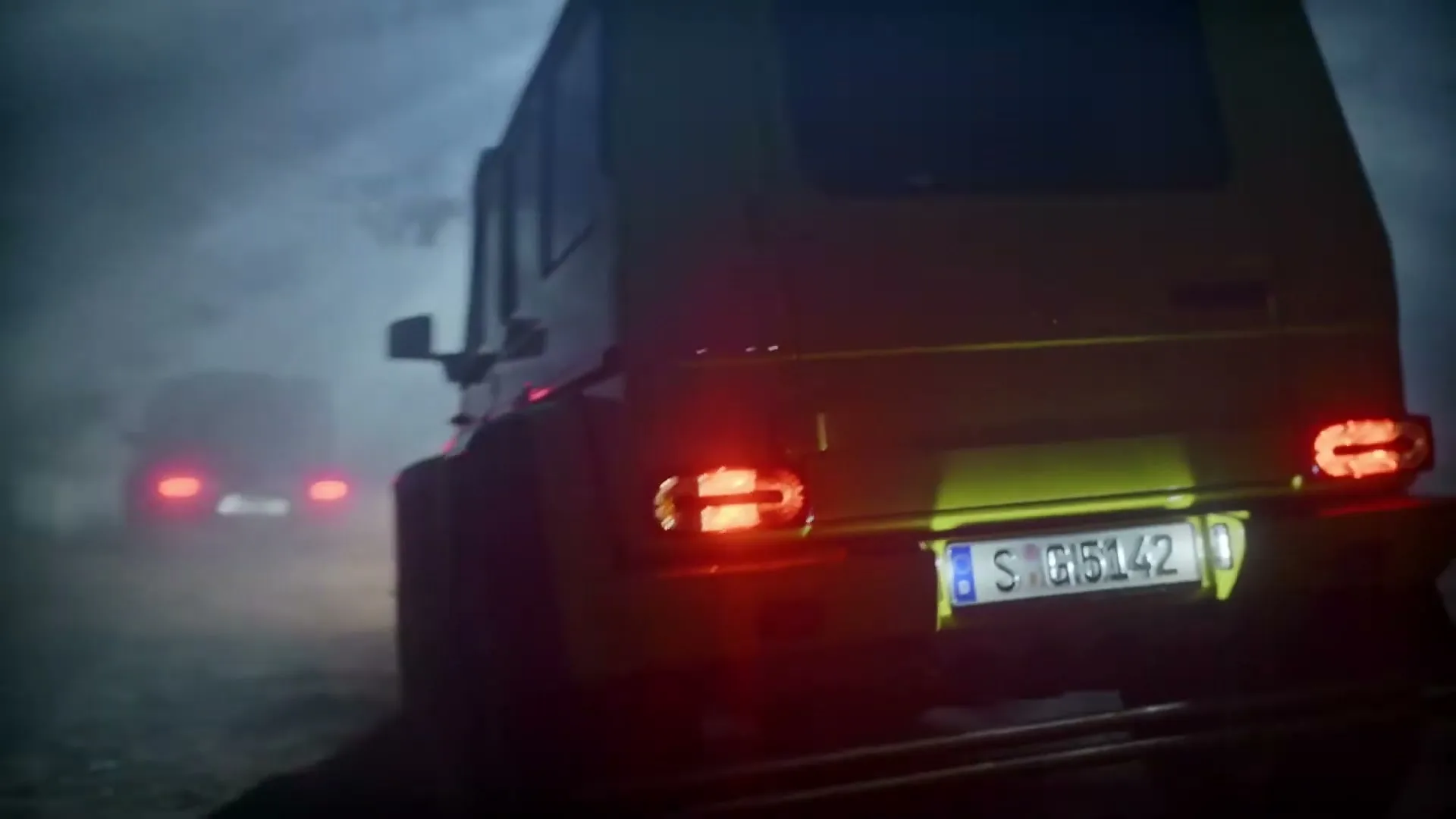 A yellow Mercedes-Benz G-Class drives away from the camera on a dark, foggy road, with another G-Class visible in the distance.