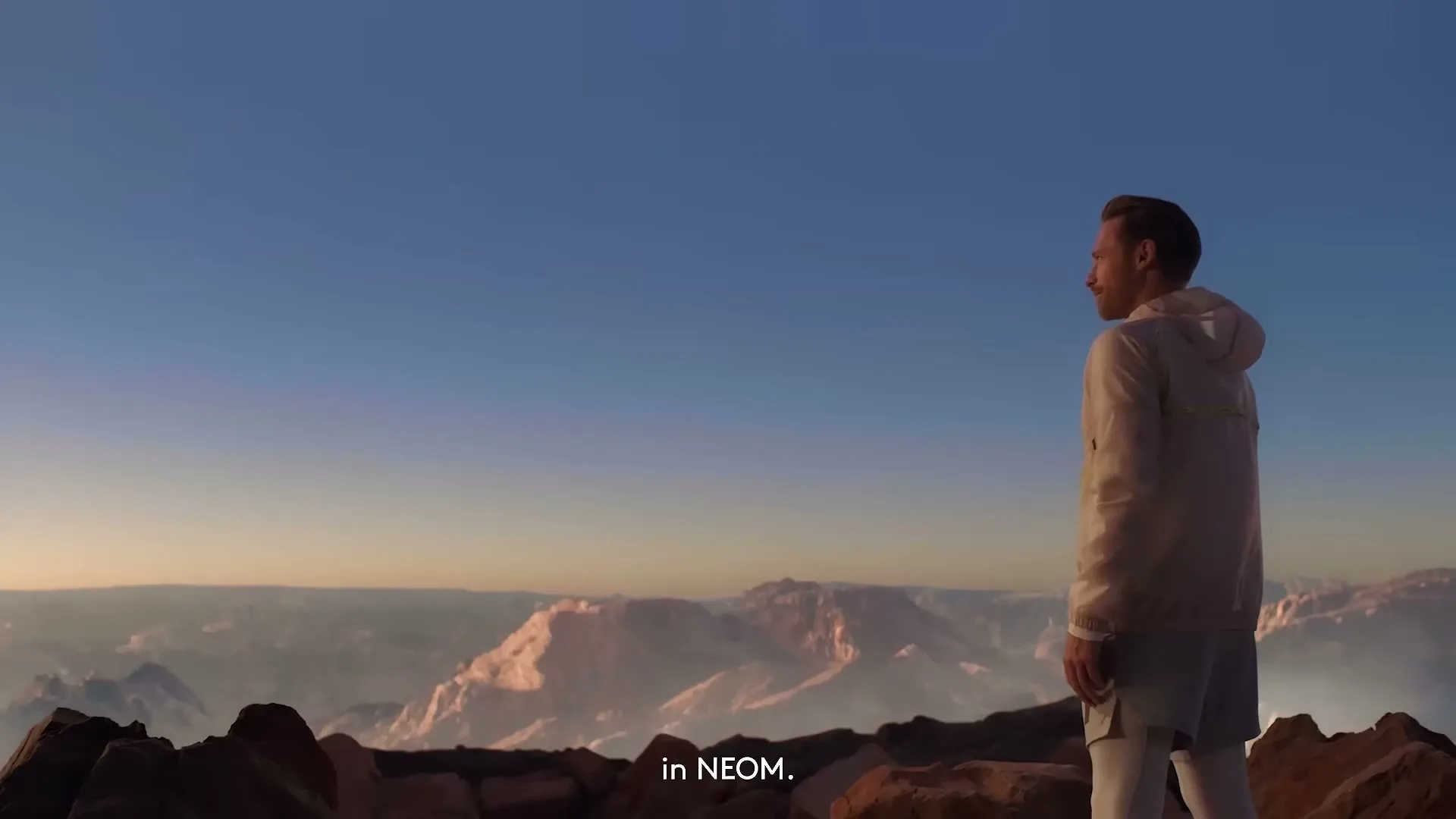 A man in athletic wear stands on a rocky mountain peak, looking out at a vast mountain range under a clear sky at golden hour.
