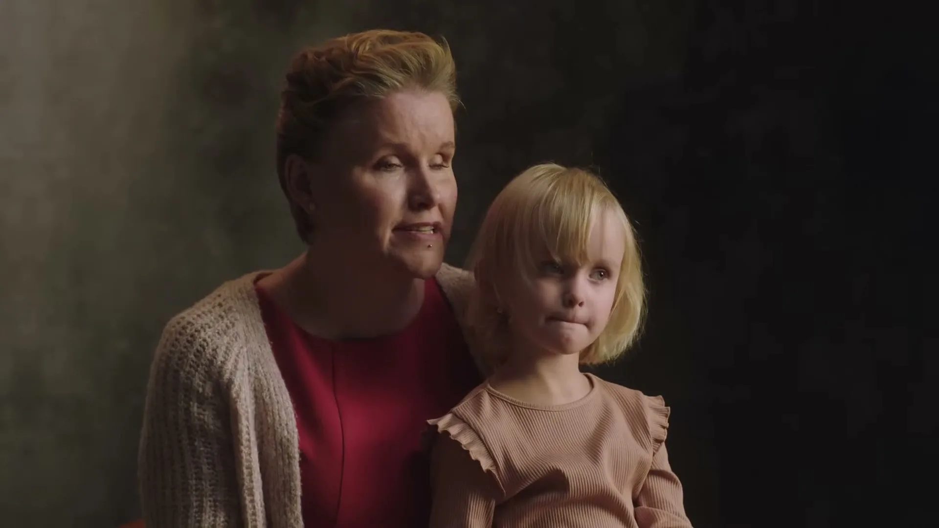 A woman with blonde hair in a red dress and a beige cardigan holds a young blonde girl in a light brown top, both looking forward with neutral expressions against a dark, smoky background.