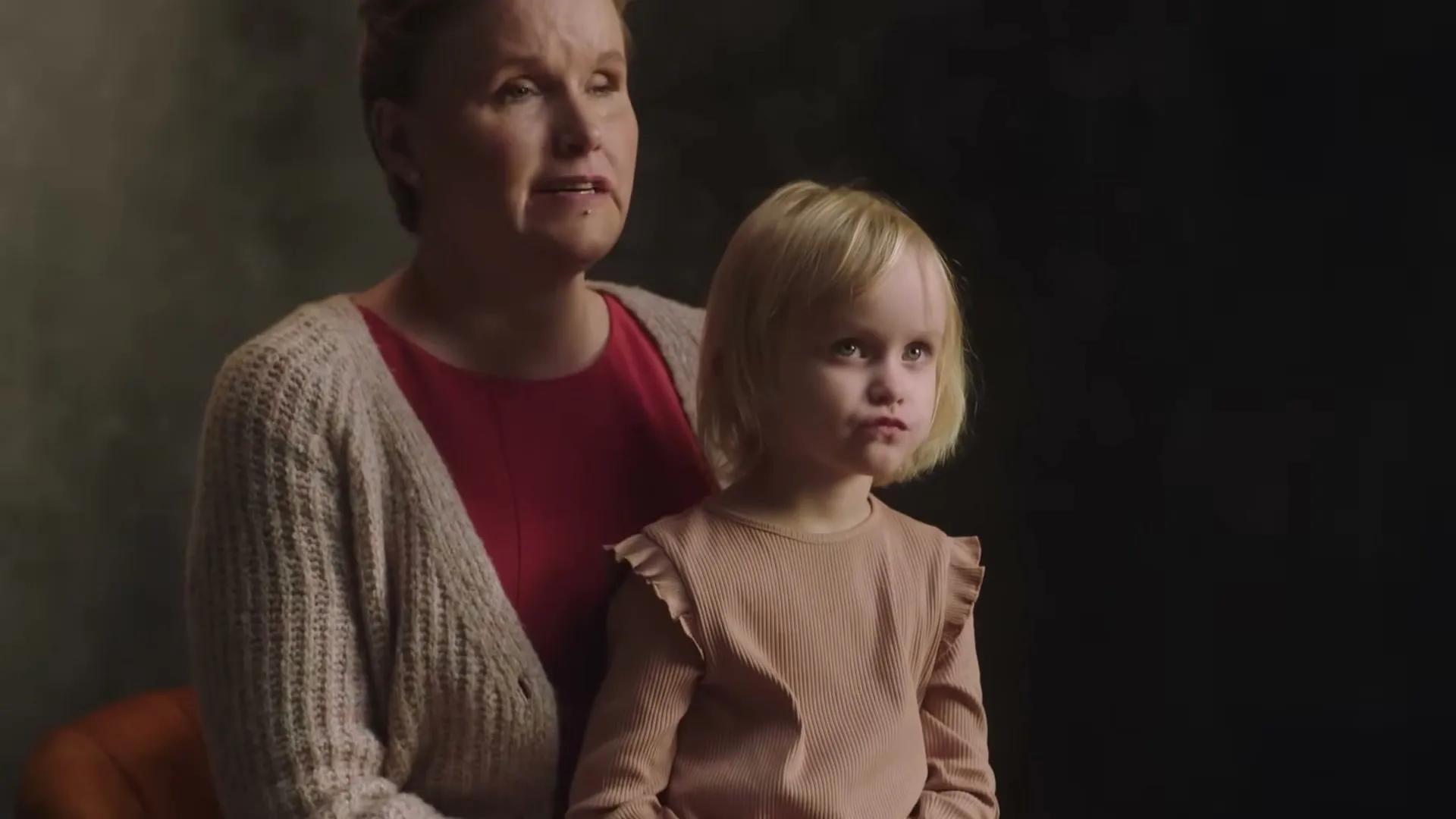 A blonde woman in her 40s, wearing a red top and beige cardigan, holds a young blonde girl in a peach top on her lap, both looking off-camera in a studio.