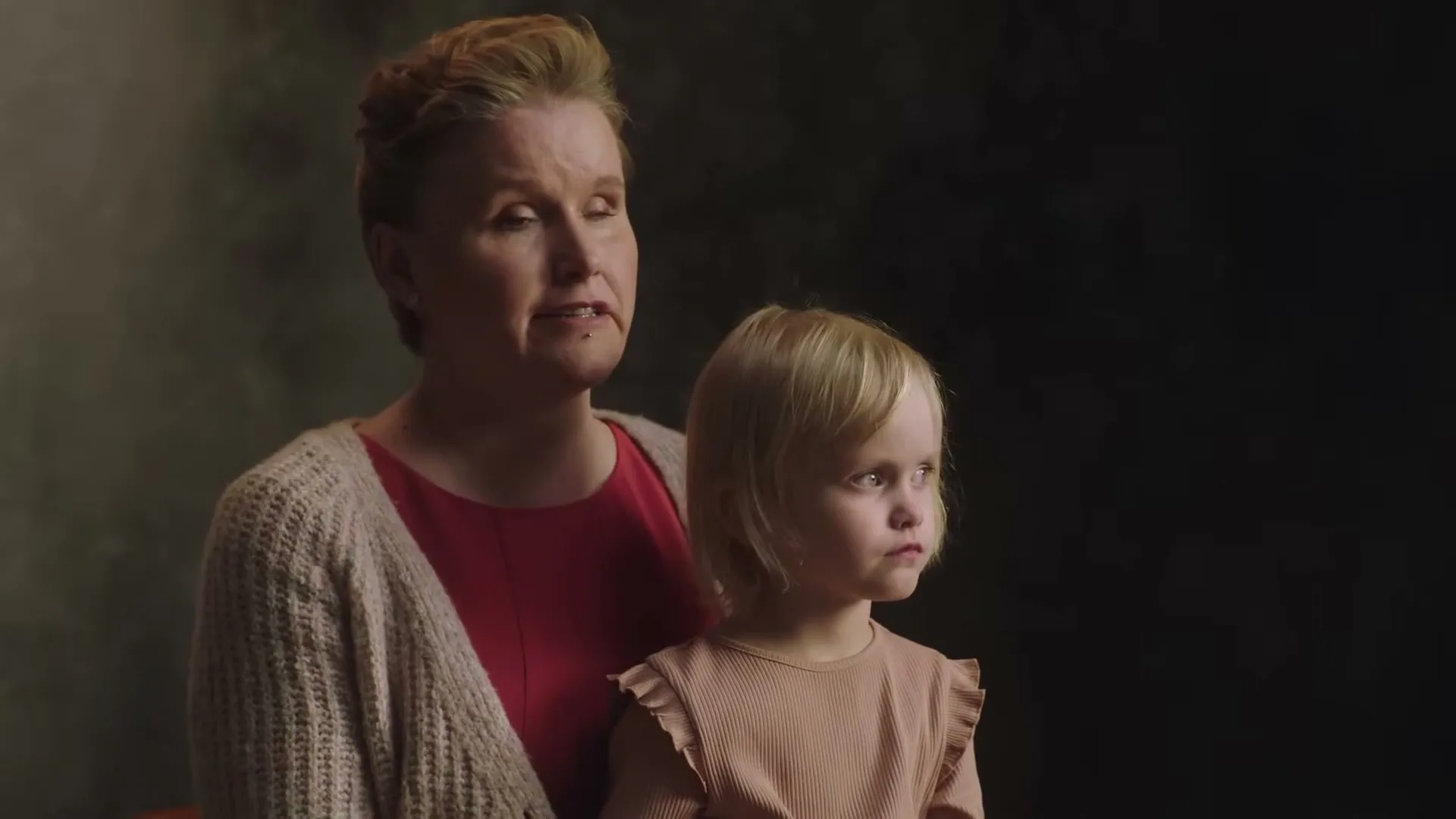 A woman in her 40s, wearing a red top and beige cardigan, holds a young blonde girl in a peach top, both looking off-camera in a studio.