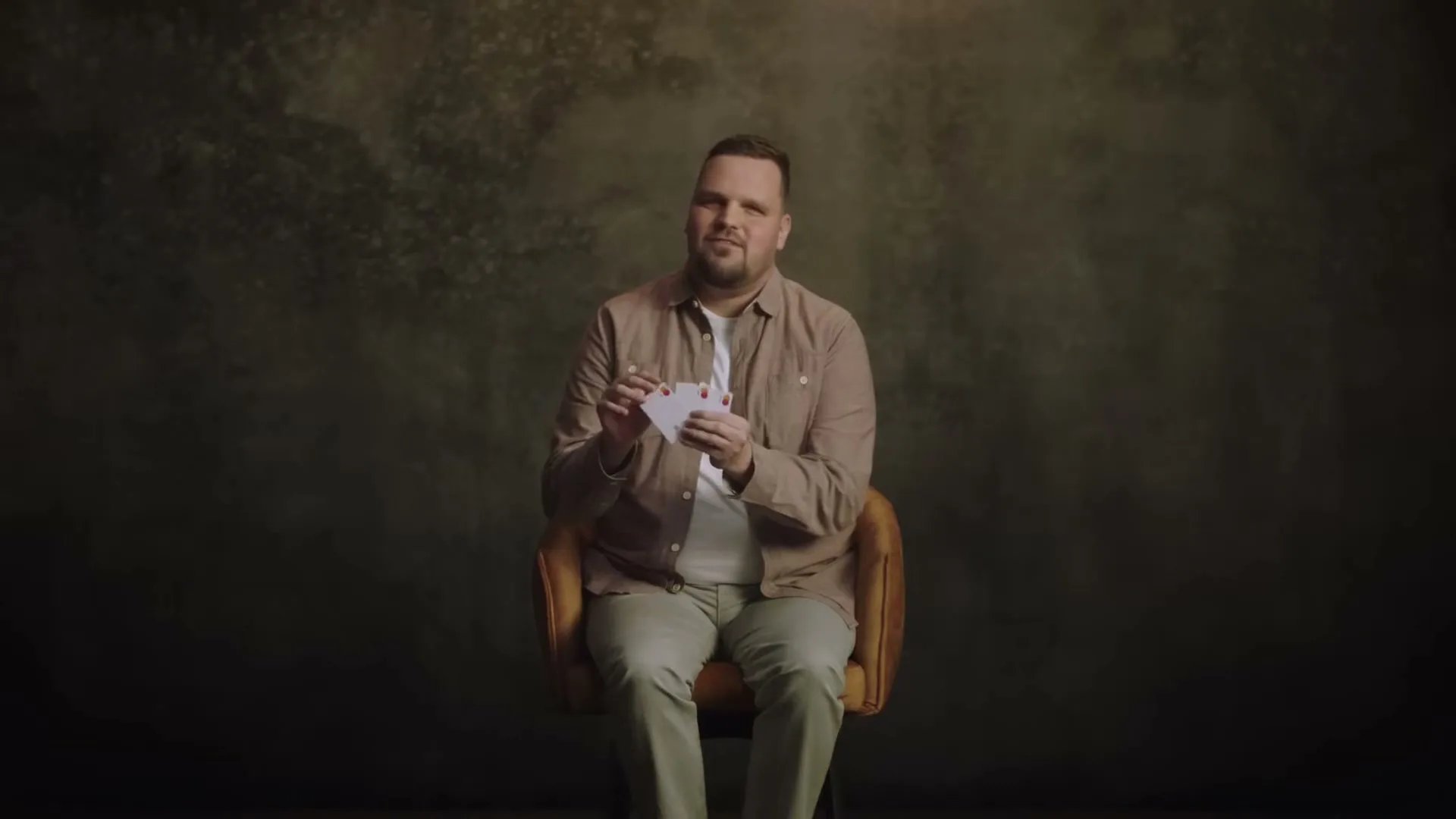 A man in his 30s, wearing a beige shirt and white t-shirt, sits in a chair, holding and looking at cards in a studio setting.