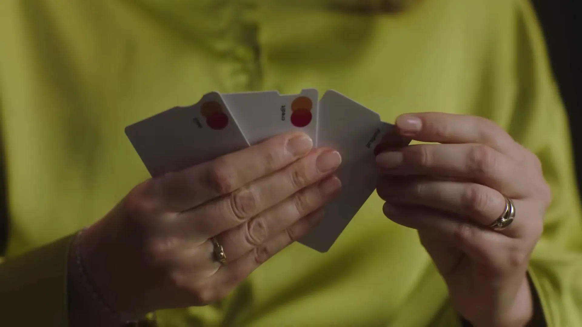 A woman's hands, wearing rings, hold and fan out three white Mastercard credit cards, revealing their distinct notched edges against a dark background.