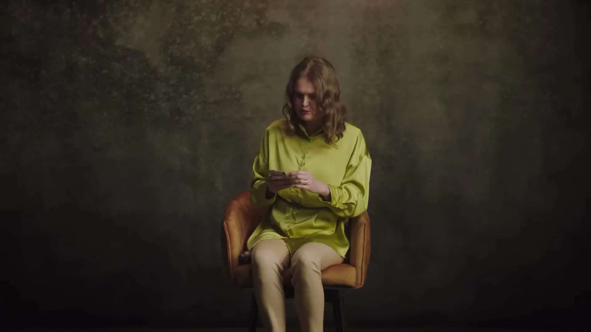 A young woman with long brown hair, wearing a lime green shirt and beige pants, sits in a brown chair, examining several cards in her hands against a dark, textured background.