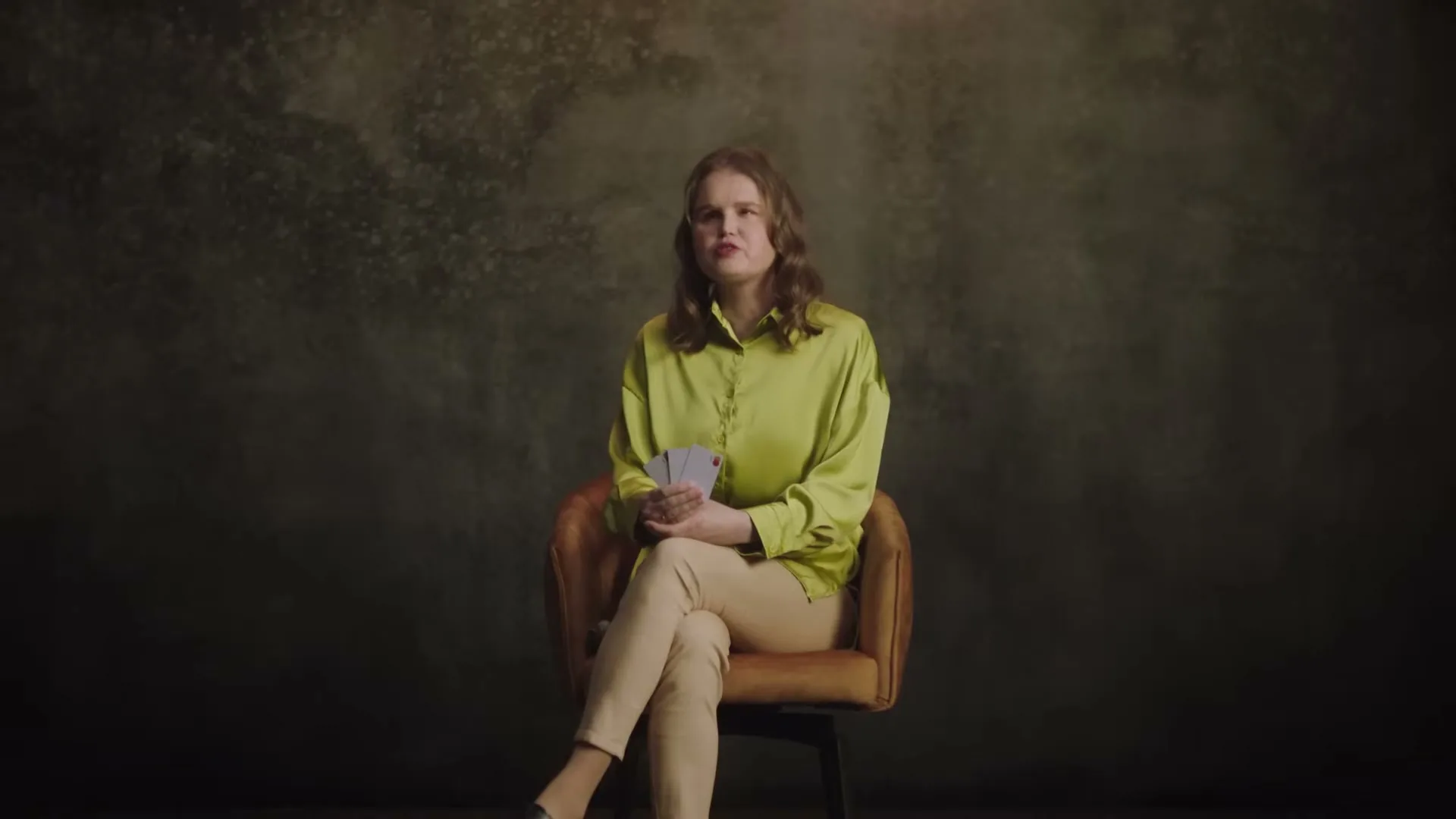 A young woman with light brown hair, wearing a lime green shirt and beige pants, sits on a brown chair, holding cards, and talking in a studio.