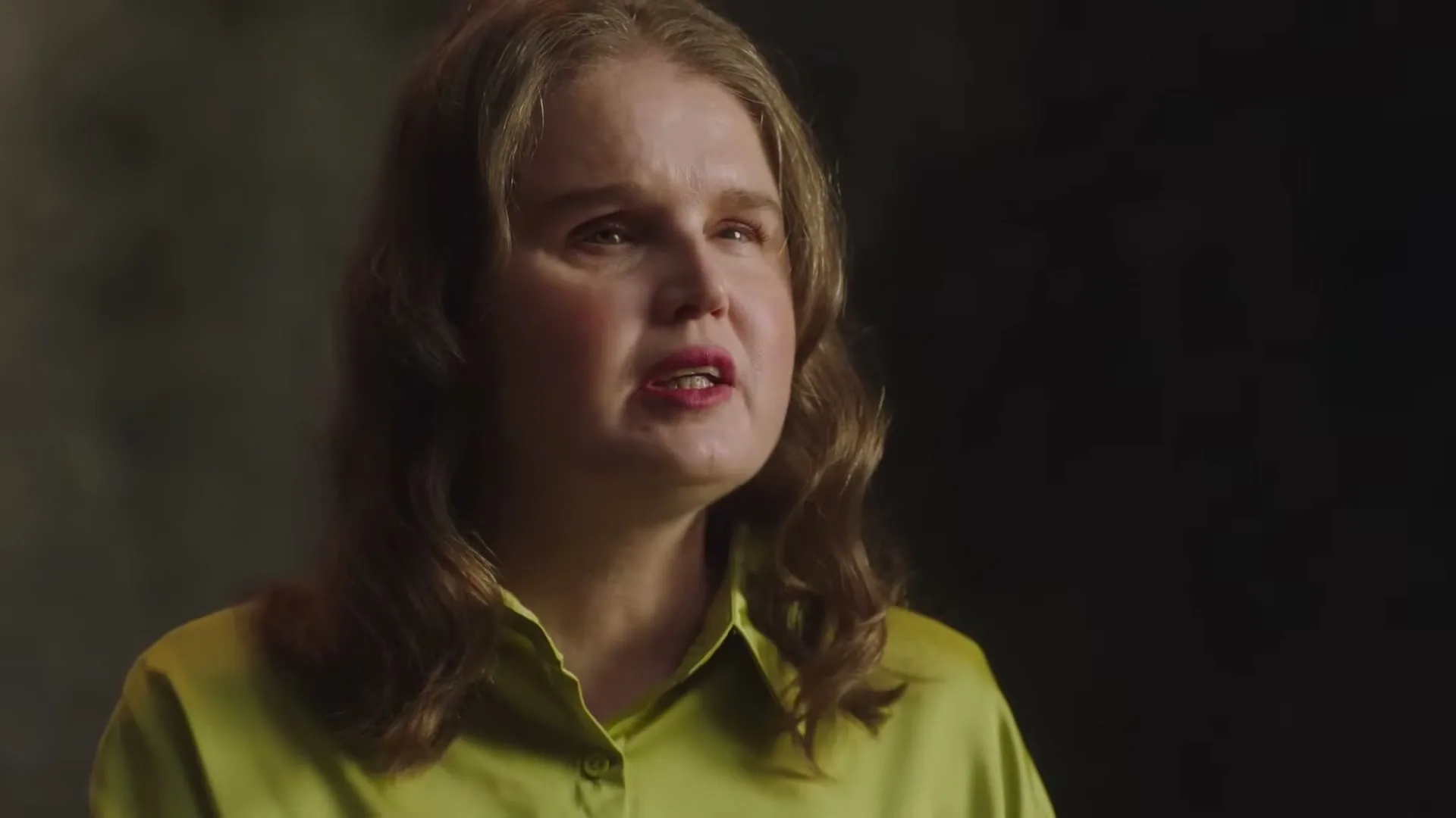 A woman with long, wavy brown hair and a bright green shirt speaks directly to the camera in a studio setting.