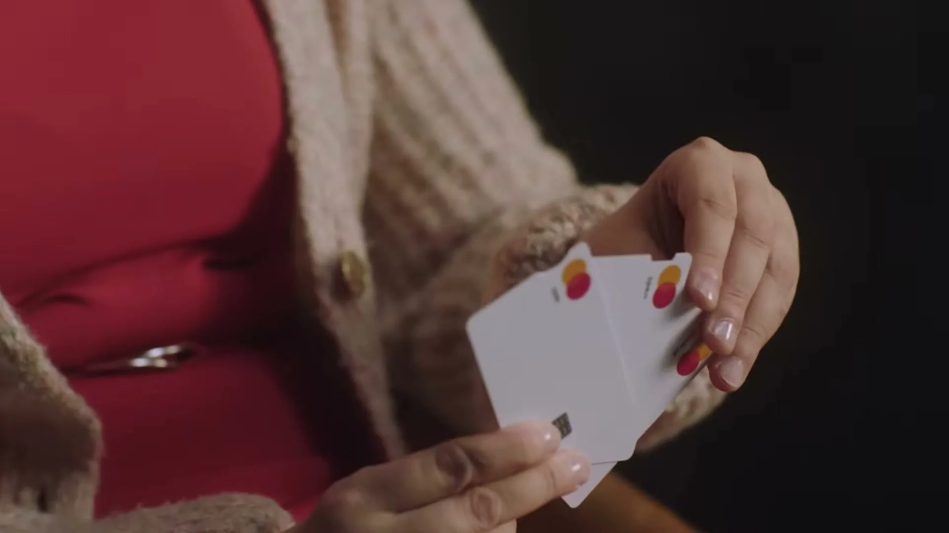 A woman in a red dress and beige cardigan holds up three white Mastercard credit cards, showing their distinct notches, against a dark background.