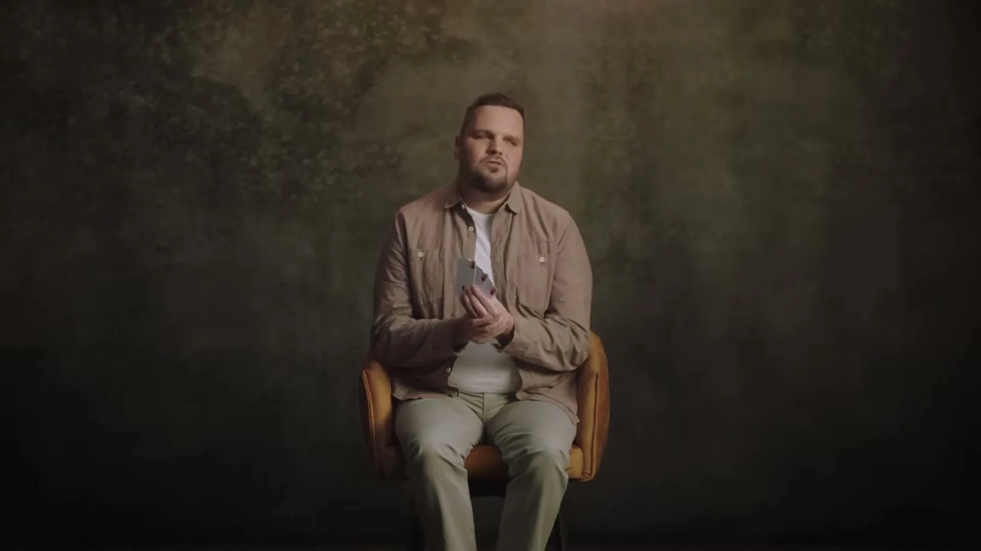 A man in his 30s, wearing a beige shirt and white t-shirt, sits in a chair, holding a Mastercard Touch Card in his hands, against a dark, textured background.