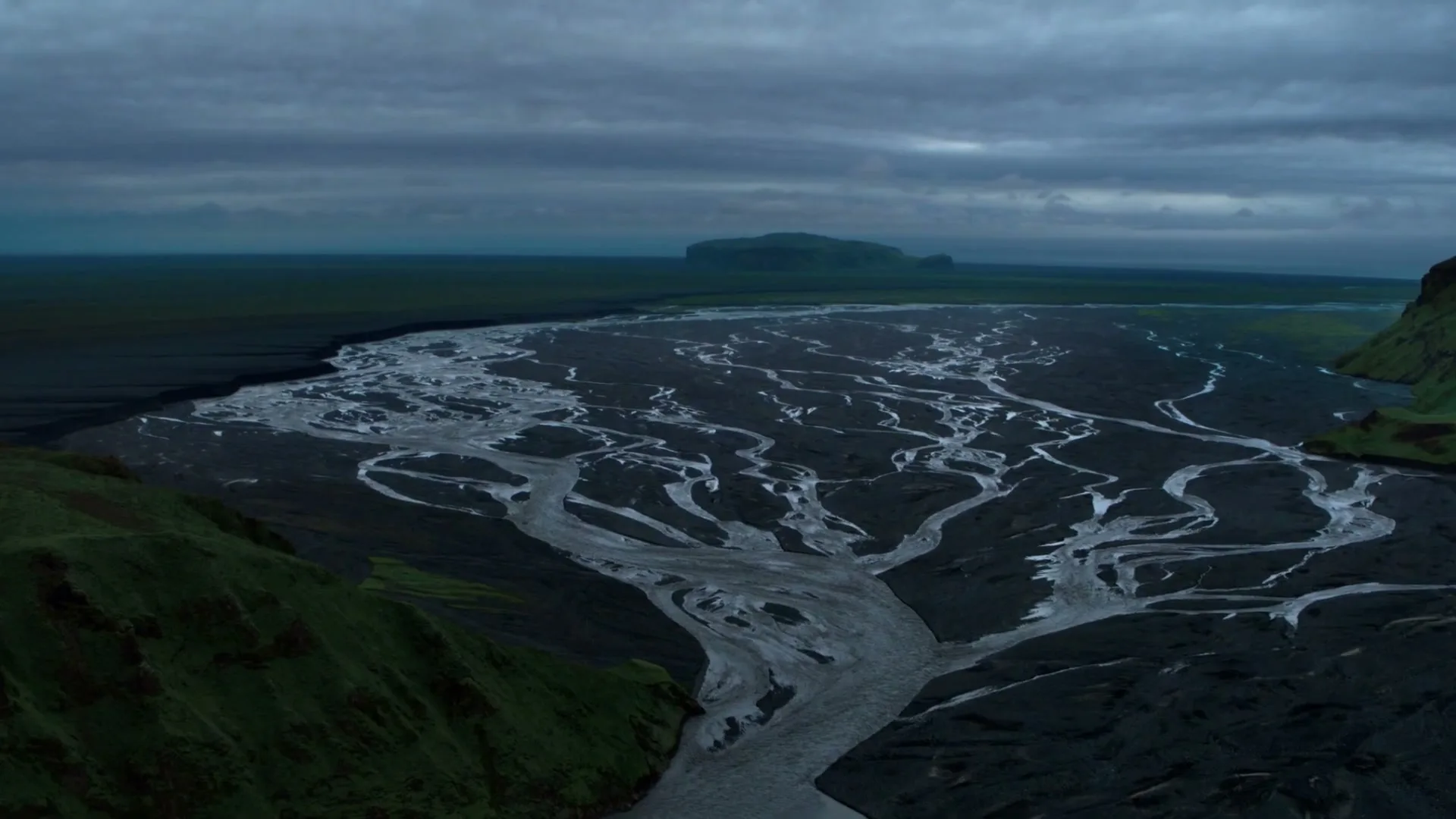 An aerial view shows a vast, dark landscape with a braided river system flowing through it under a cloudy, overcast sky.