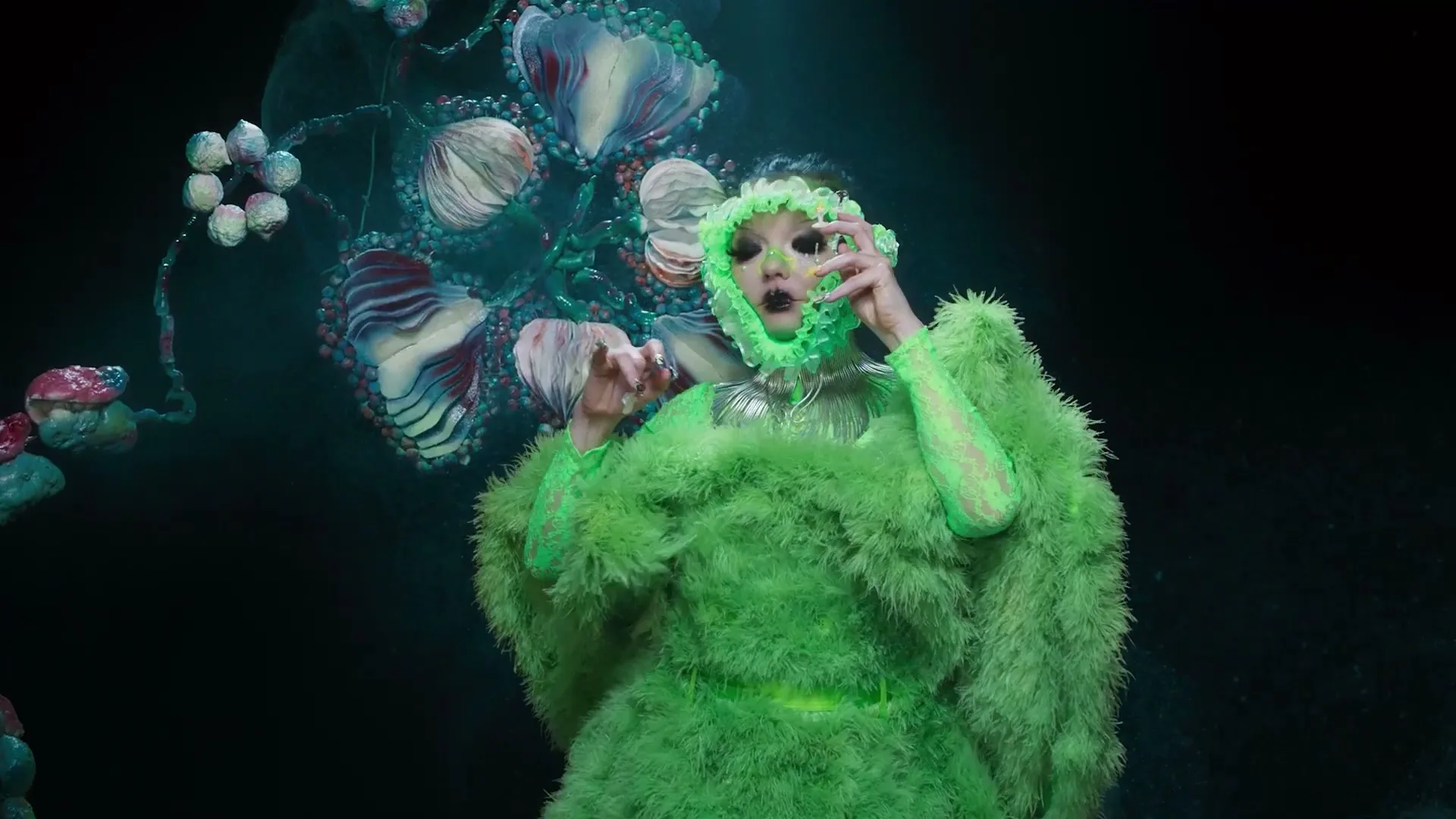 Björk, dressed in a vibrant green, feathery costume and an elaborate headpiece, performs with expressive hand gestures against a dark, fantastical background.