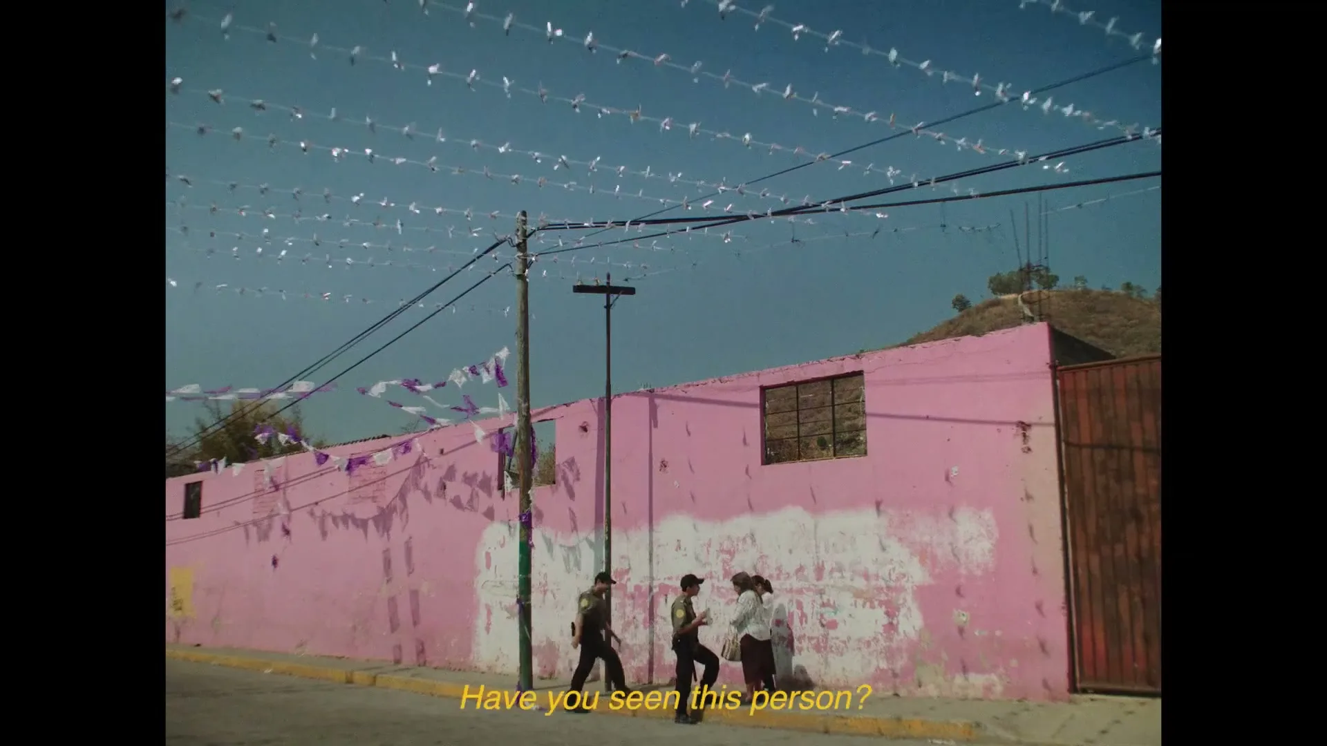 Two men in uniform and a woman in a white shirt stand on a sidewalk next to a pink wall with white paint peeling off, under a clear blue sky with string lights.