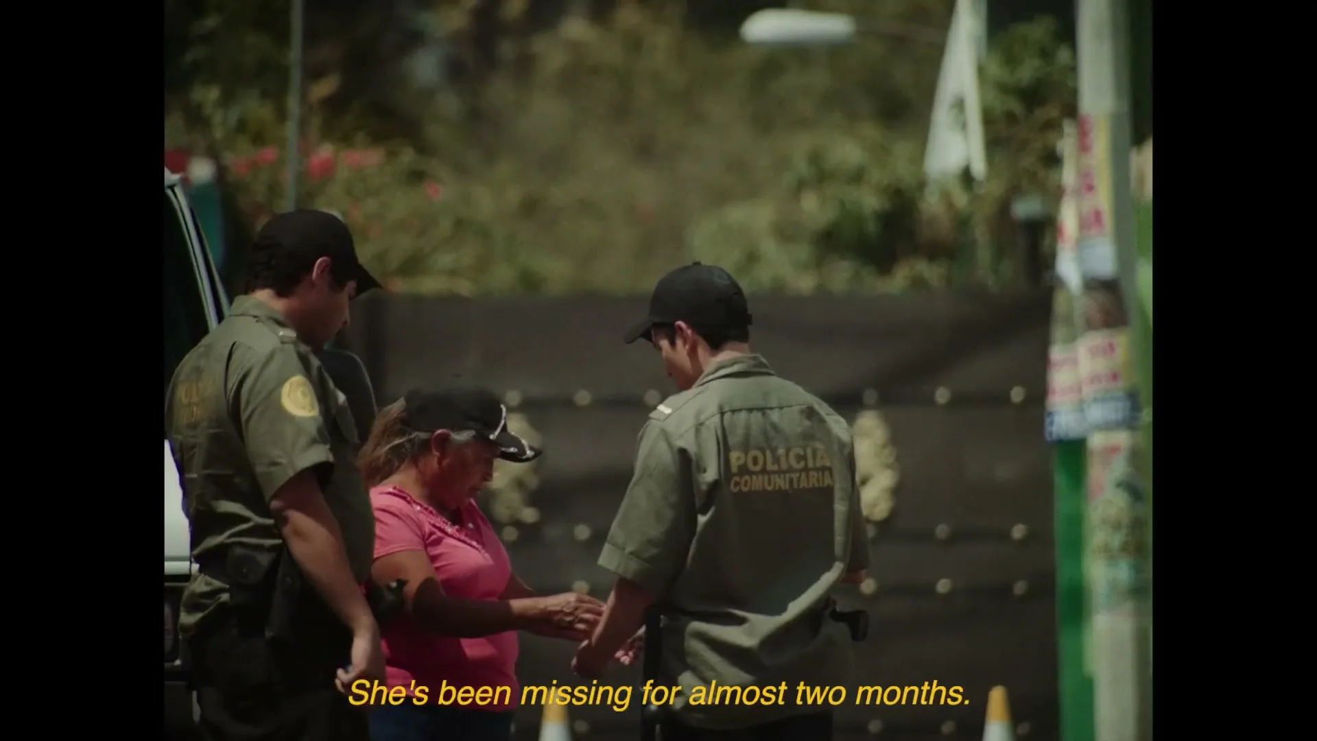 Two male police officers in green uniforms and a woman in a pink shirt and black cap stand outside, talking, with a dark gate and foliage in the background.