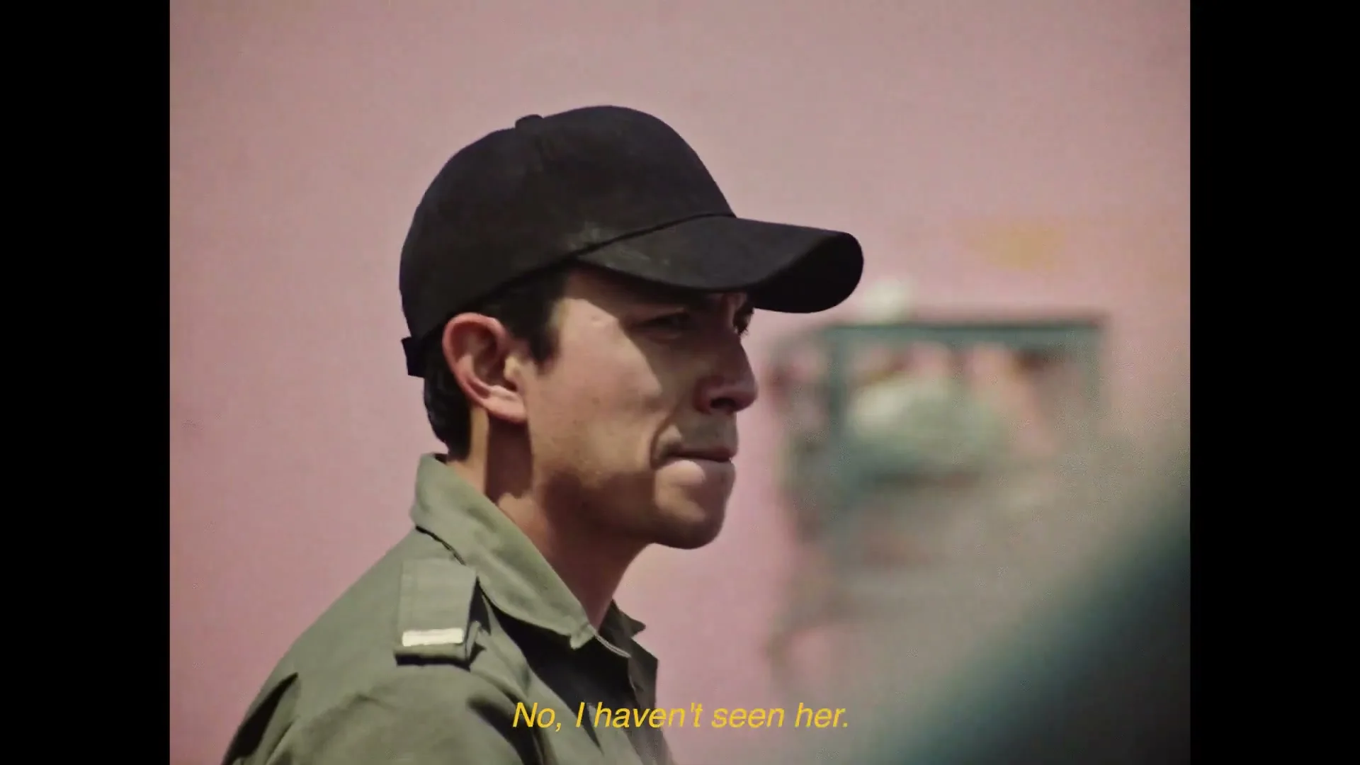 A male police officer in a black cap and green uniform speaks to someone off-camera, with a pink wall in the background.