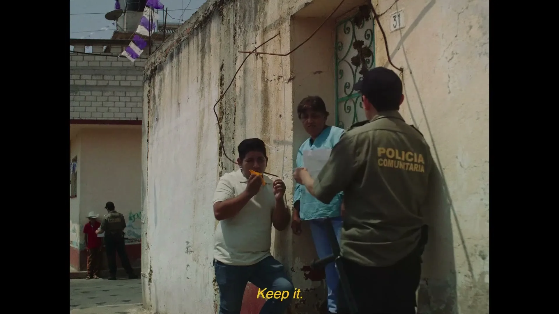 A community police officer in uniform talks to a young boy holding a yellow flower and a woman in a doorway on a sunny day in a rural village.