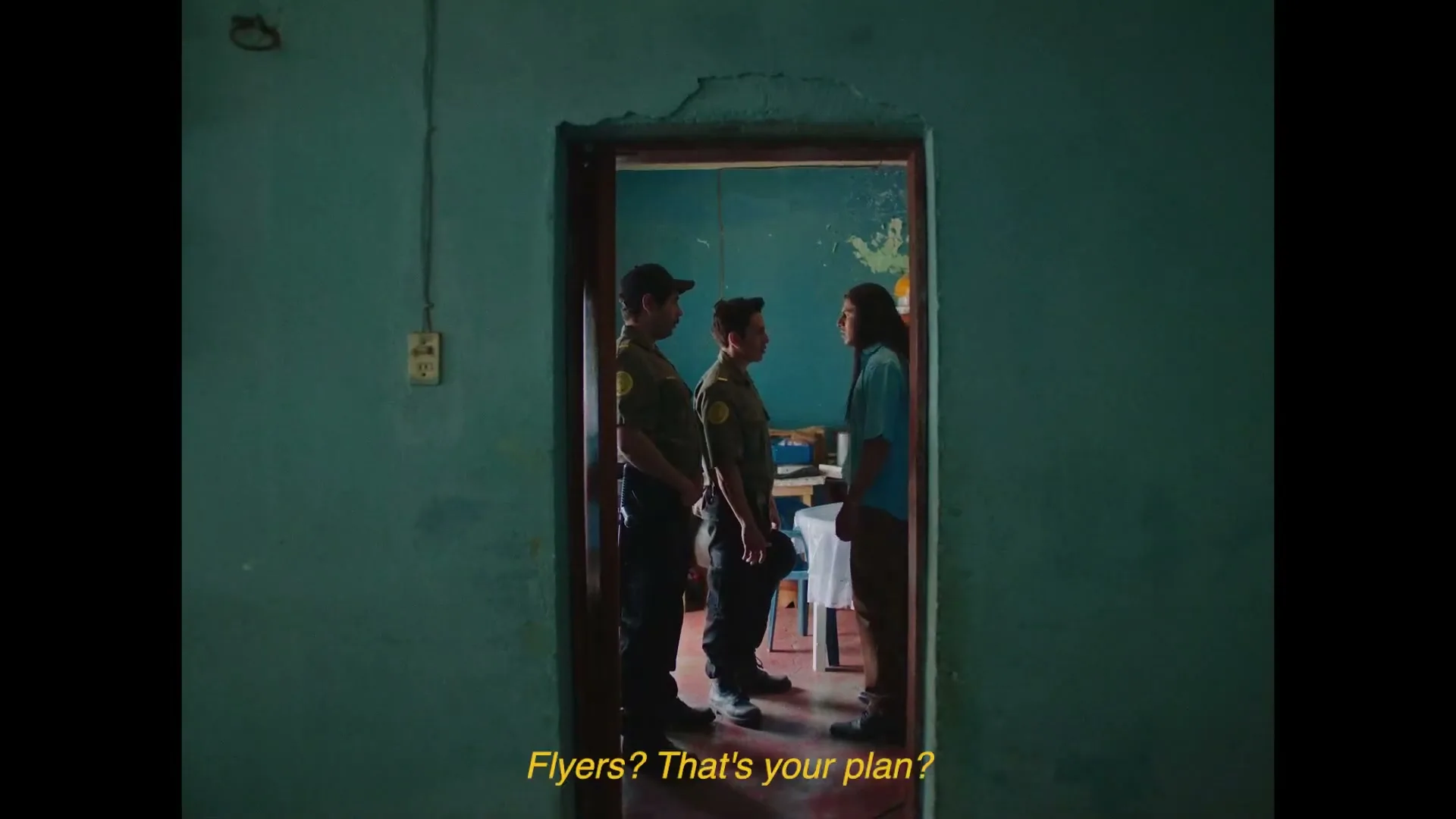 Three men, two in uniform and one in a blue shirt, stand in a doorway, arguing intensely in a dimly lit room.