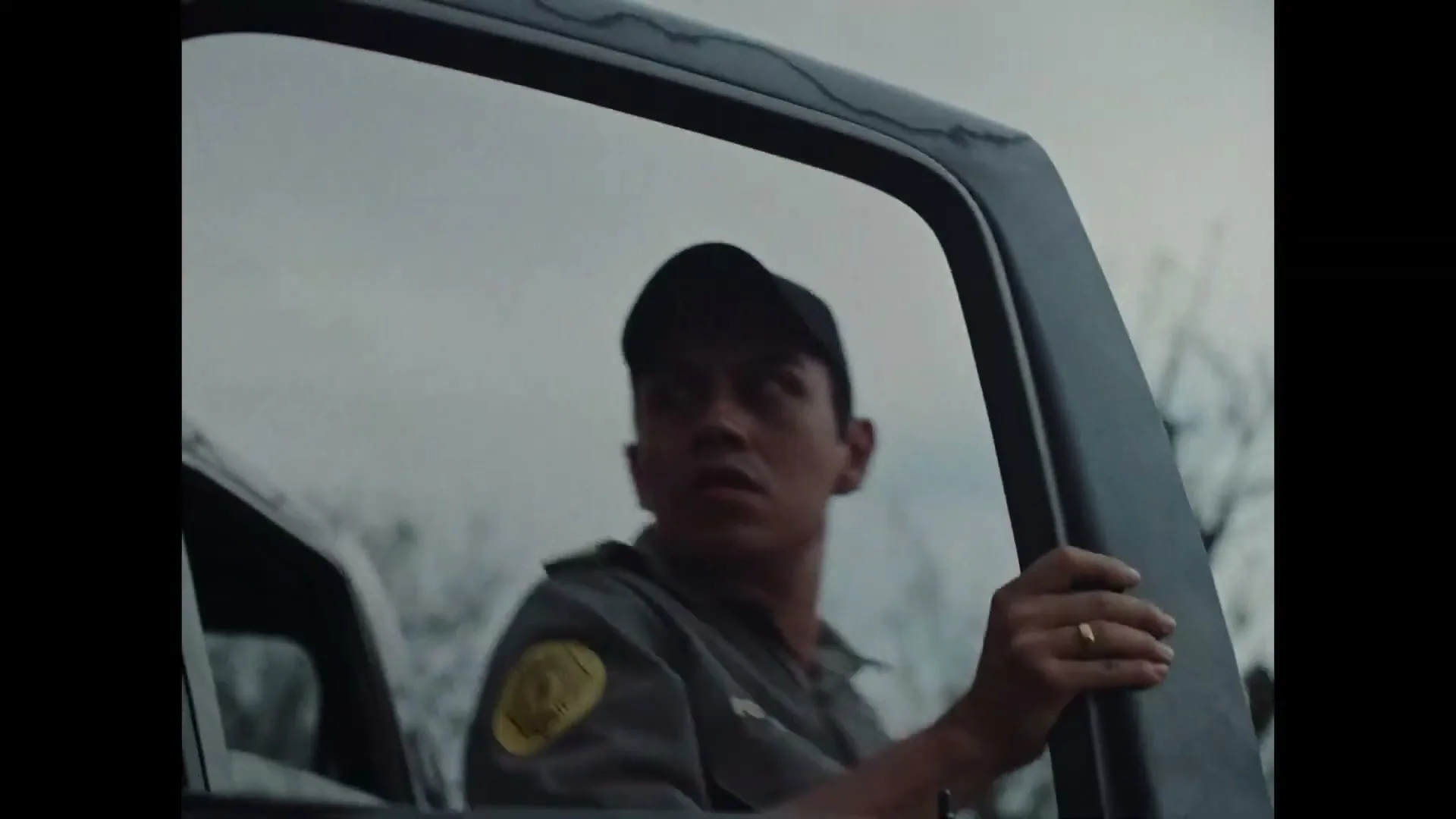 A male police officer in a cap and uniform exits a police car, looking around with a concerned expression under a cloudy sky.