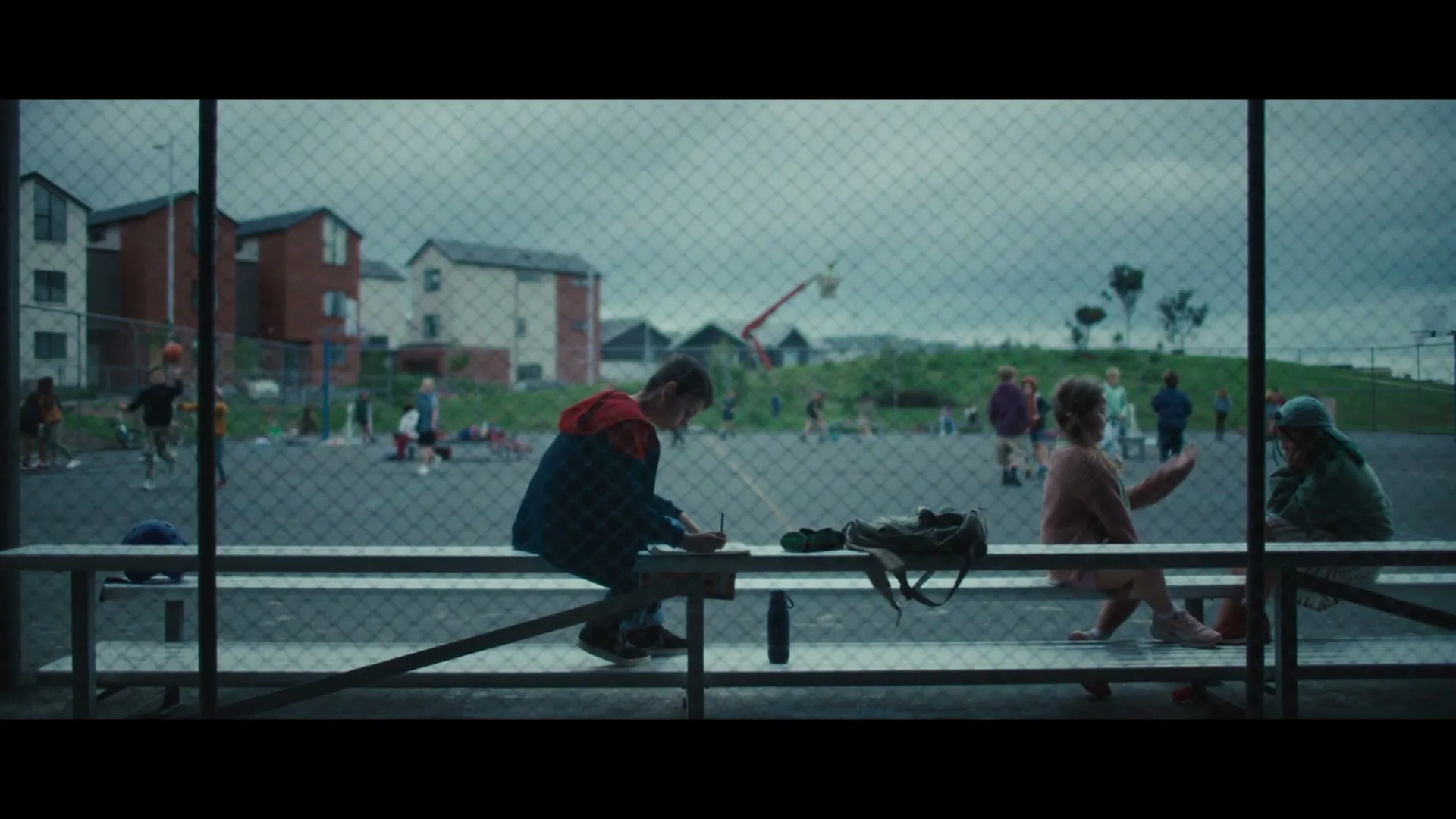 A young boy sits on a bench, writing in a notebook, while two girls sit nearby, all framed by a chain-link fence, on a cloudy day at an outdoor court.