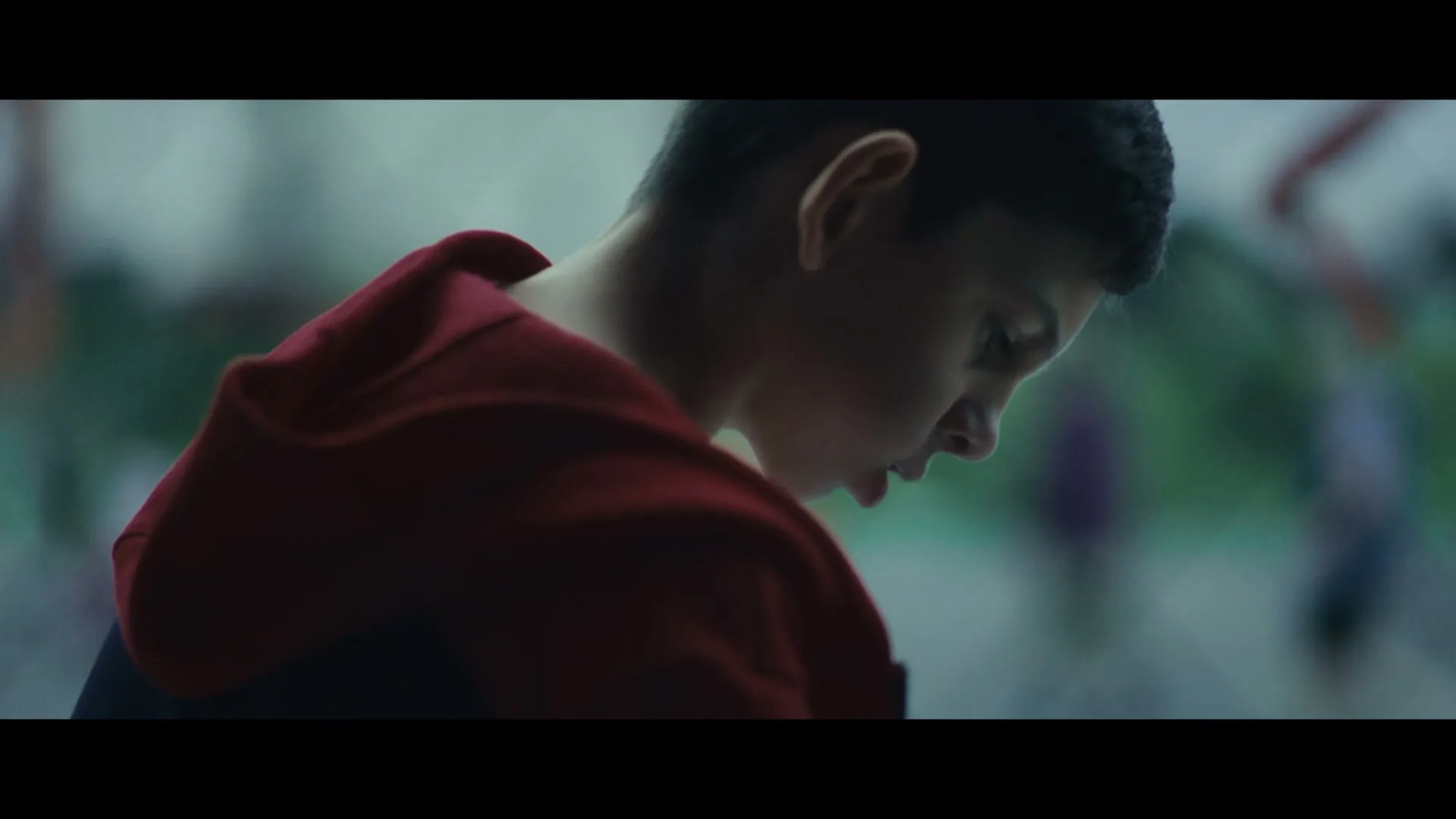 A young boy with dark hair, wearing a red hoodie, looks down with a somber expression in an interior setting, possibly a classroom.