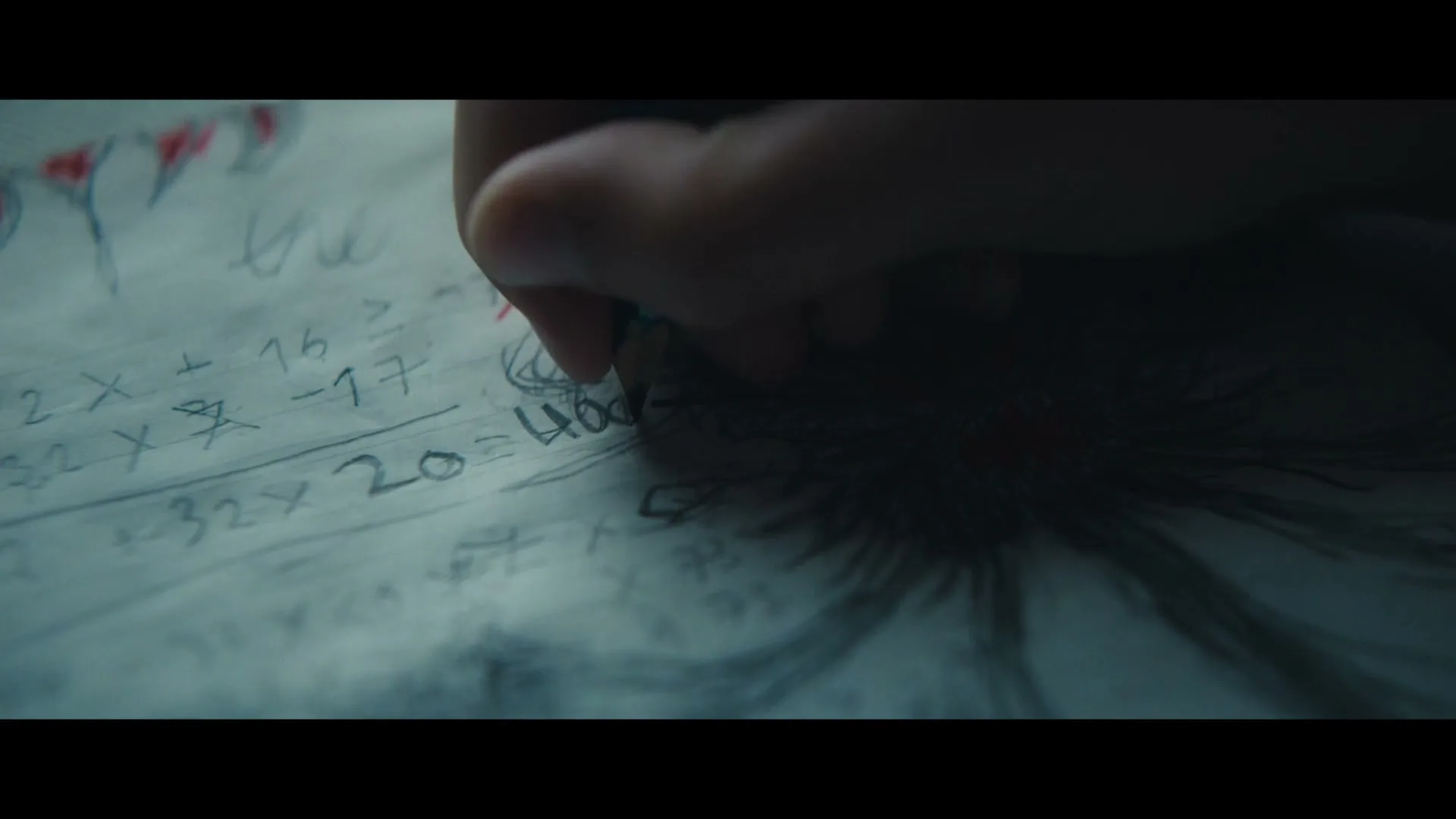 A child's hand holds a pencil, writing on a piece of paper with math equations and a monster drawing, in a dimly lit interior.