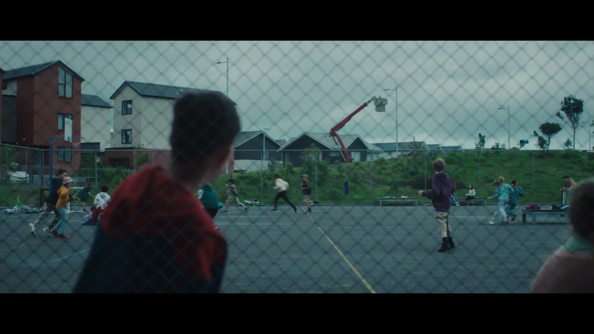 A young boy in a red hoodie watches other children playing on a basketball court, as a large, shadowy monster appears in the background.