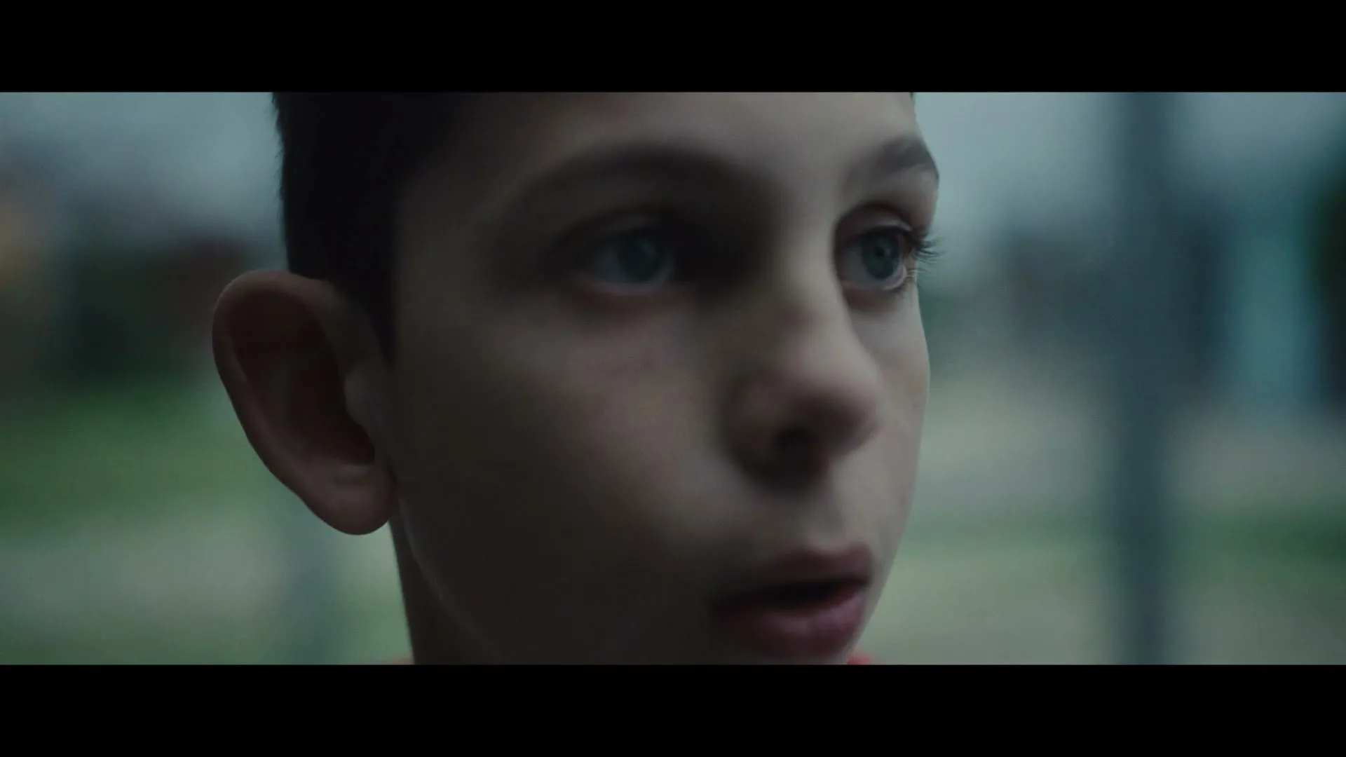 A young boy with blue eyes and dark hair looks up with a worried expression, his face partially obscured by shadow.