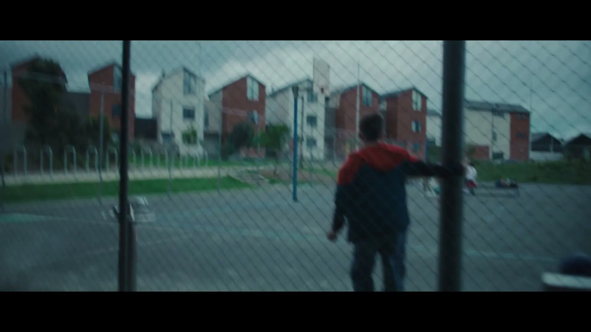 A boy in a red and blue jacket sits on a bench, watching other children play on a basketball court, as a monster appears in the background.